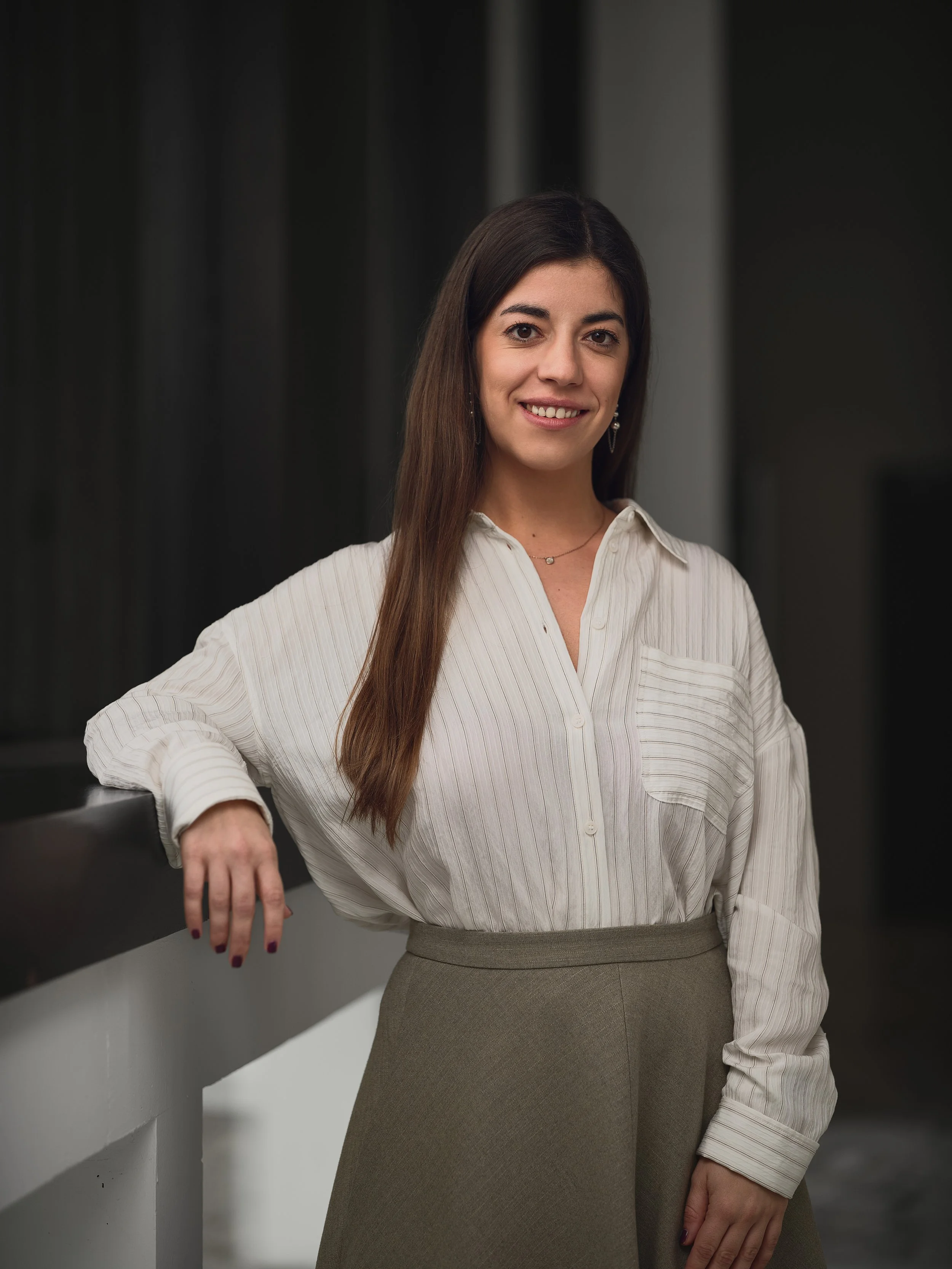A woman with long brown hair, wearing a beige blouse and a gray skirt, standing next to a railing in an indoor setting, smiling at the camera.
