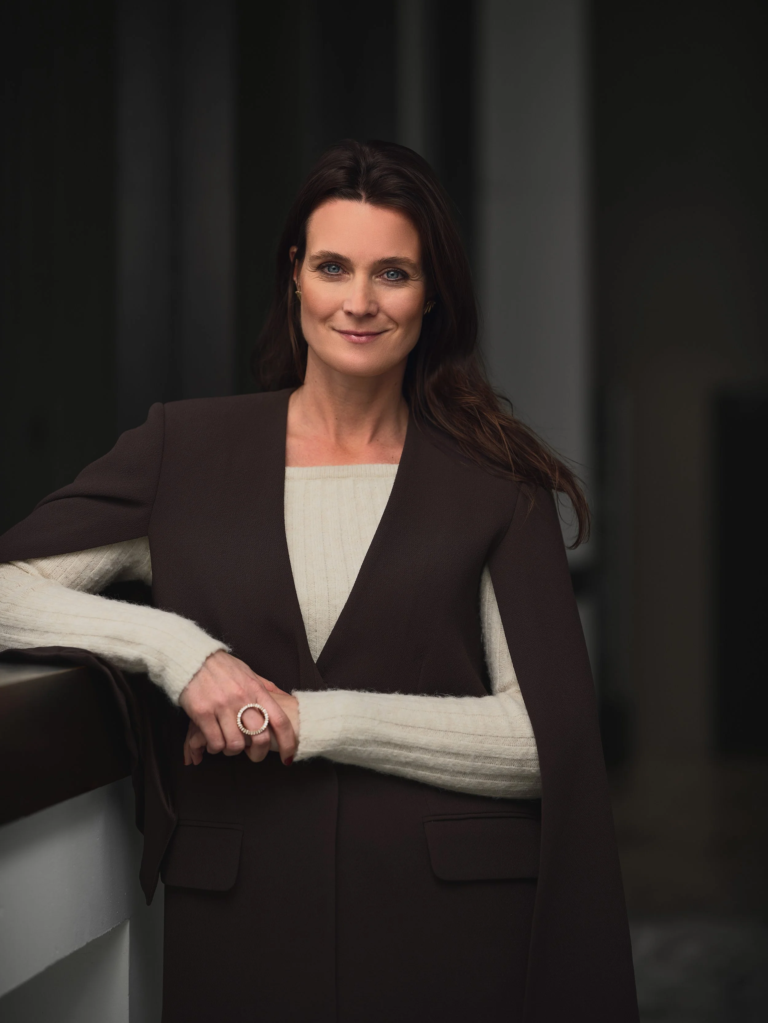 A woman with dark hair and blue eyes, wearing a dark blazer over a cream-colored sweater, standing in a dimly lit corridor with dark curtains in the background.