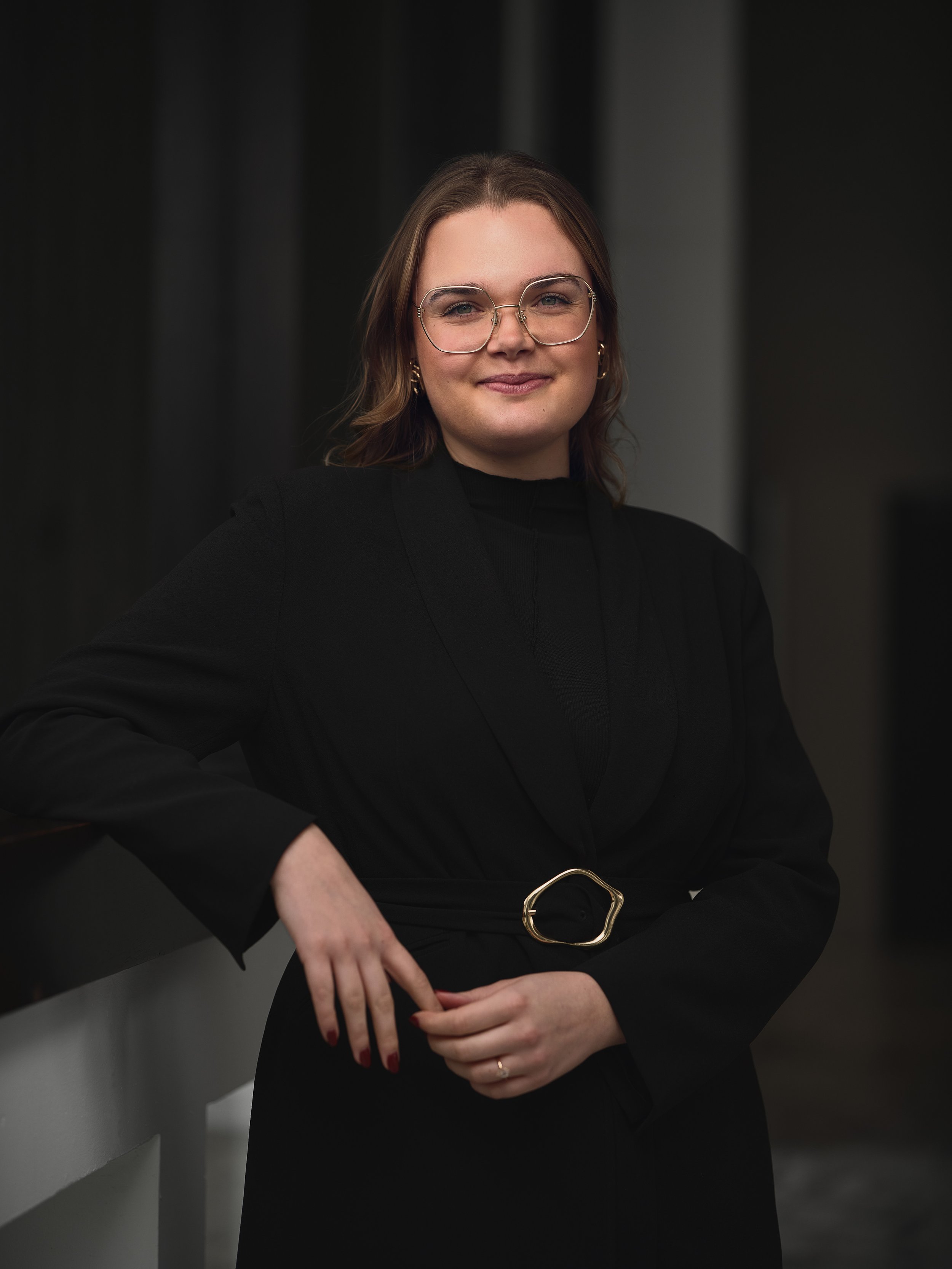 A woman with shoulder-length brown hair, wearing glasses and a black outfit with a gold belt buckle, is standing against a dark background and smiling at the camera.