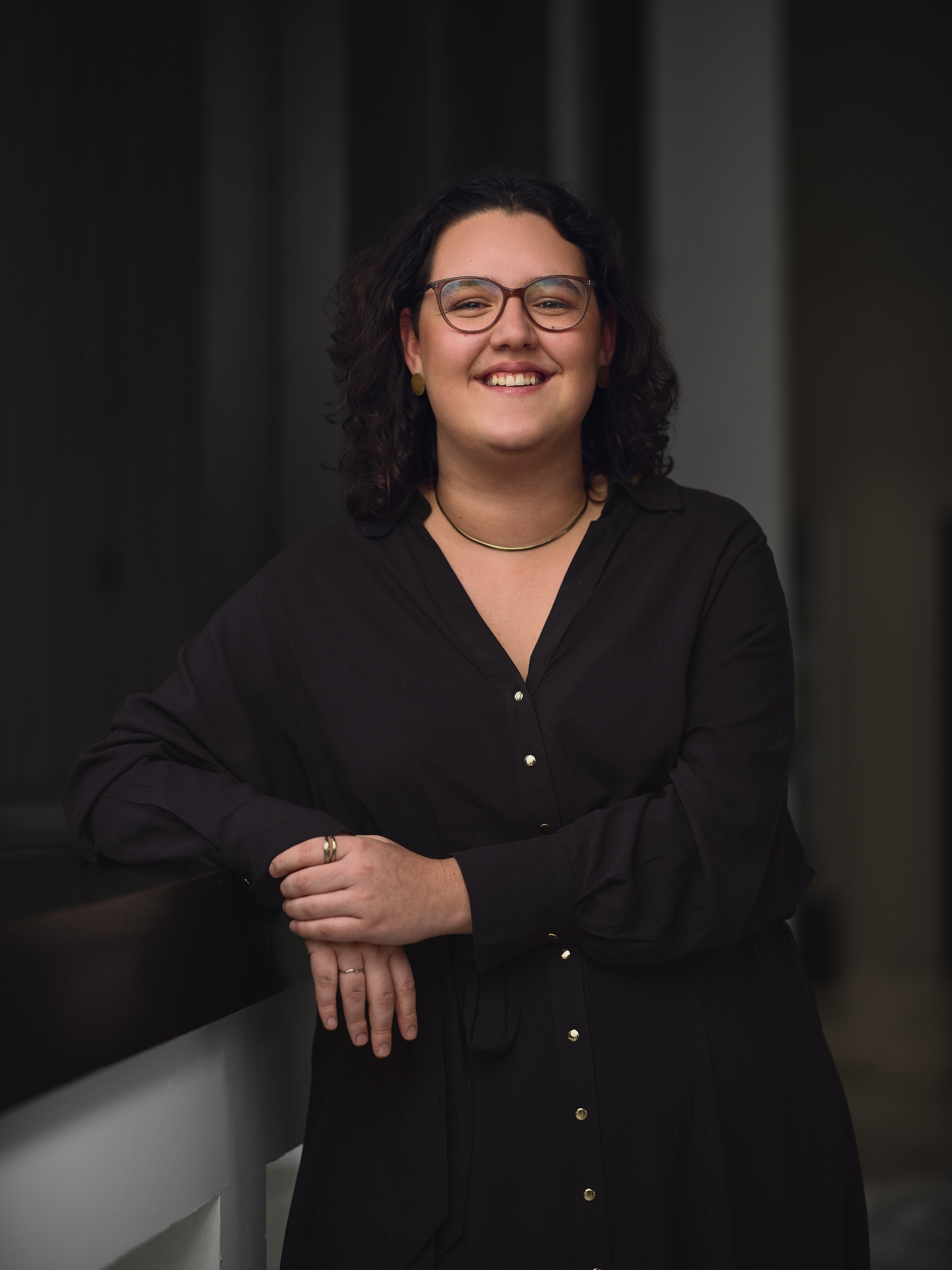 A woman with curly dark hair and glasses smiling, wearing a black blouse with gold buttons, standing against a dark, blurred background.