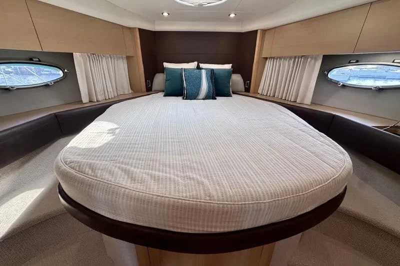 A bedroom with a large, oval bed with a striped mattress, multiple teal and white pillows, and round windows with beige curtains on each side.