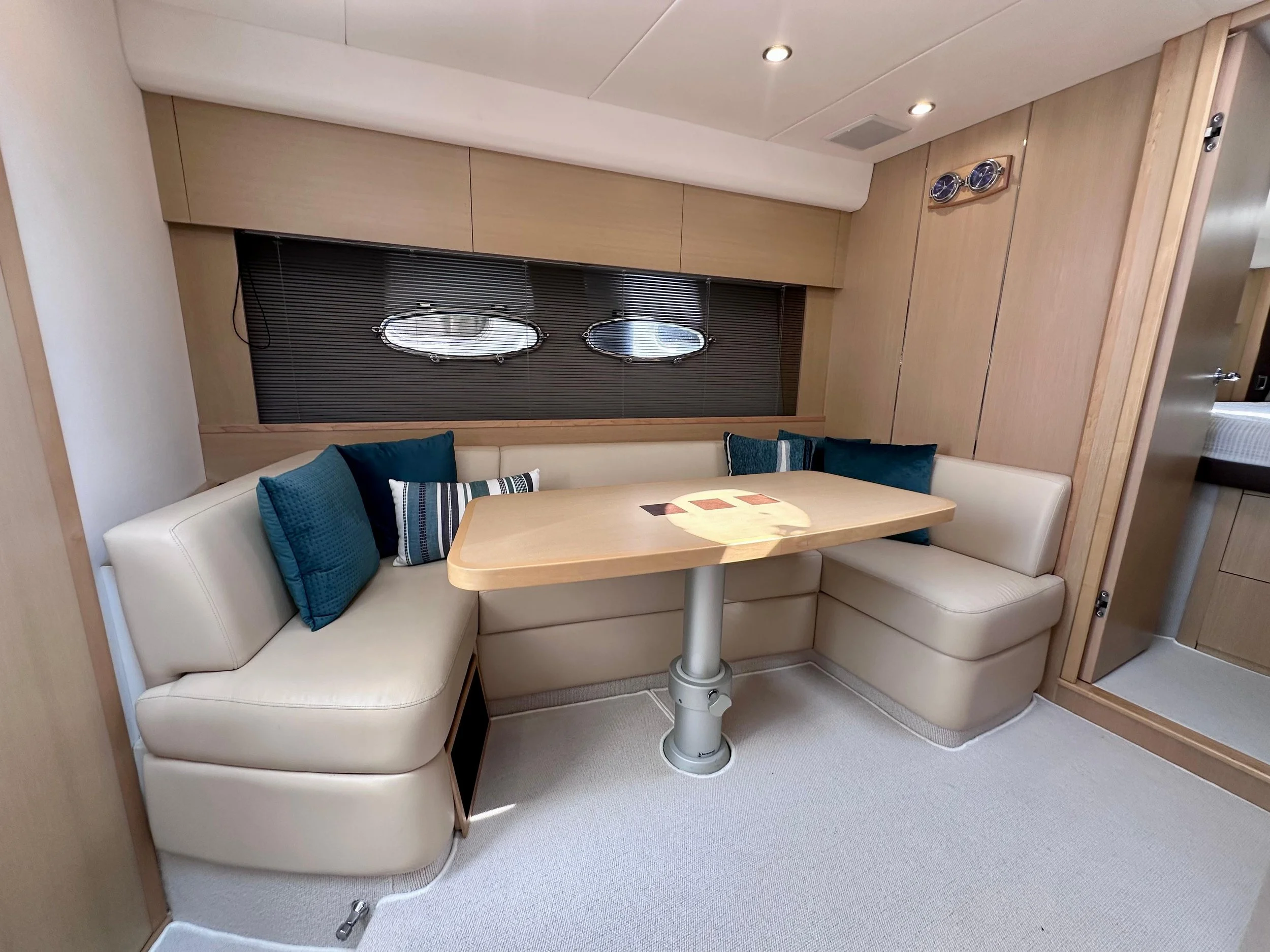 A cozy dining nook with a rounded beige leather banquette, a wooden table with a star design, and blue pillows, in a room with wood-paneled walls and small oval porthole windows.
