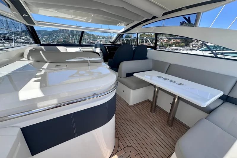 Exterior of a yacht with white and navy seating, a white table with cup holders, large panoramic windows showing a marina and mountains outside, and a curved ceiling.