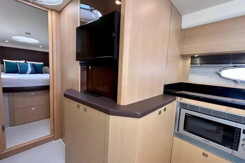 Interior of a yacht or boat with a bedroom visible through an open door, a flat-screen TV mounted on a wooden panel, and a microwave oven built into the cabinetry next to a small window.
