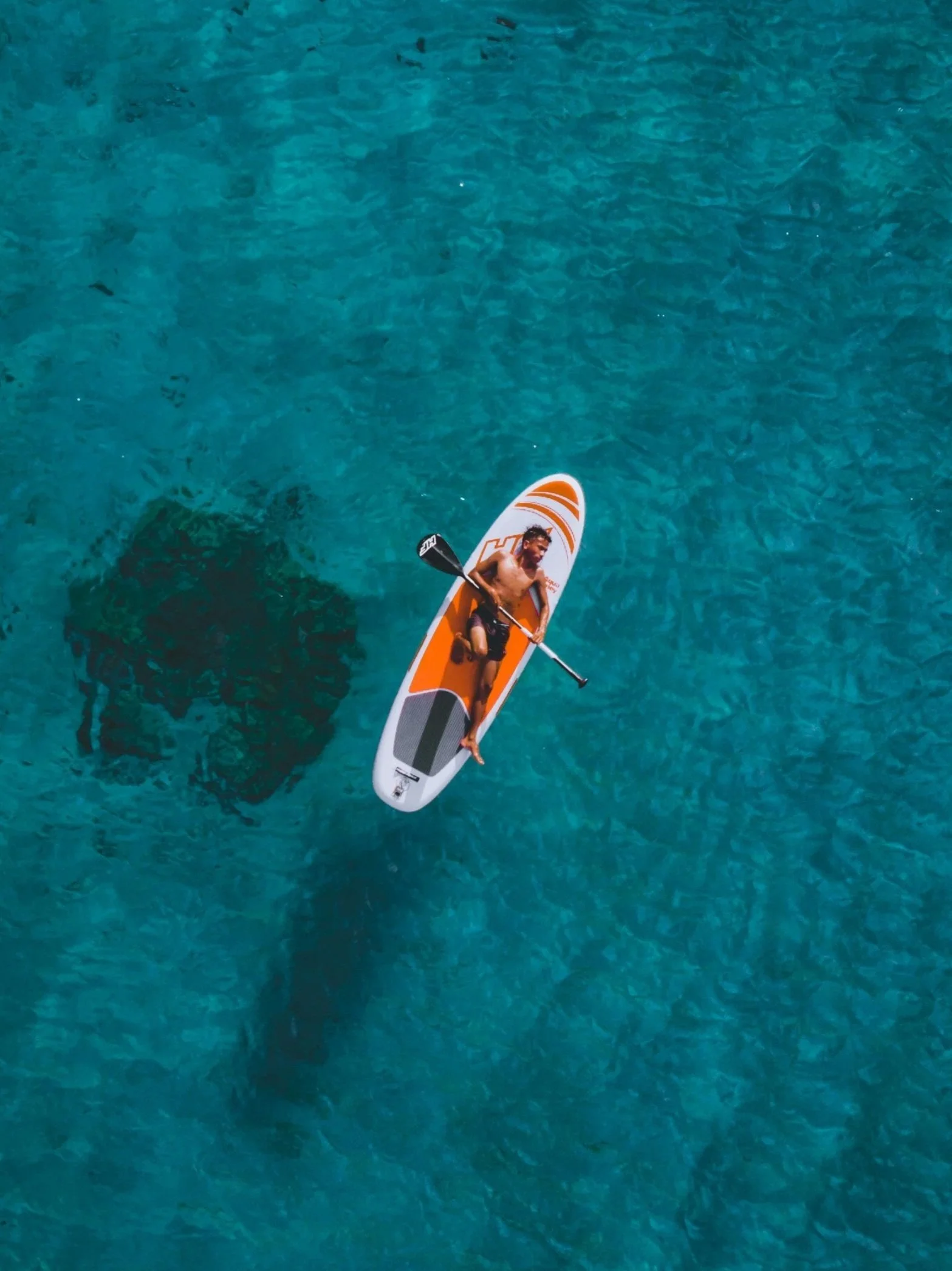 A man paddling on a paddleboard in clear blue water, with a shadow of the paddleboard visible beneath him.