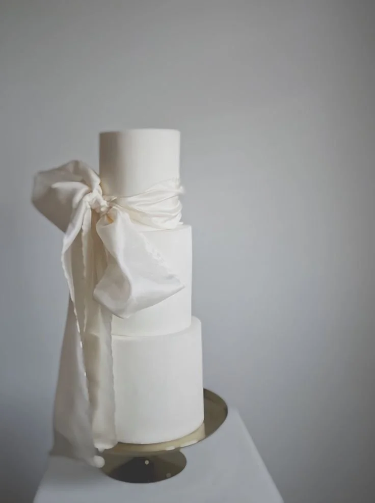 Elegant white tiered wedding cake with a large decorative bow on the side, placed on a reflective surface against a plain gray background.