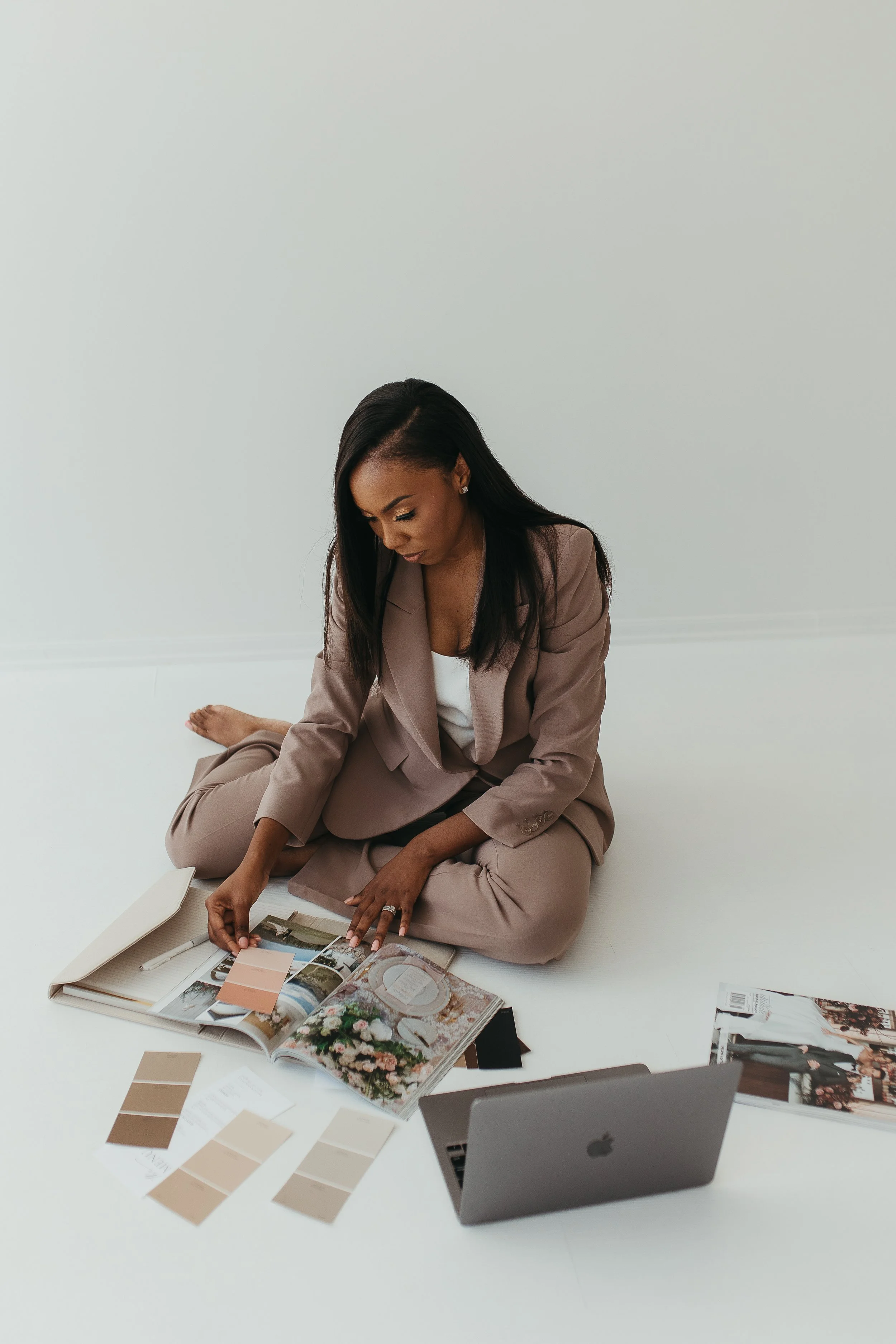 A woman dressed in a beige suit sitting on the floor, looking at and arranging multimedia design materials, including magazines, color samples, and printed photos, with a laptop nearby.