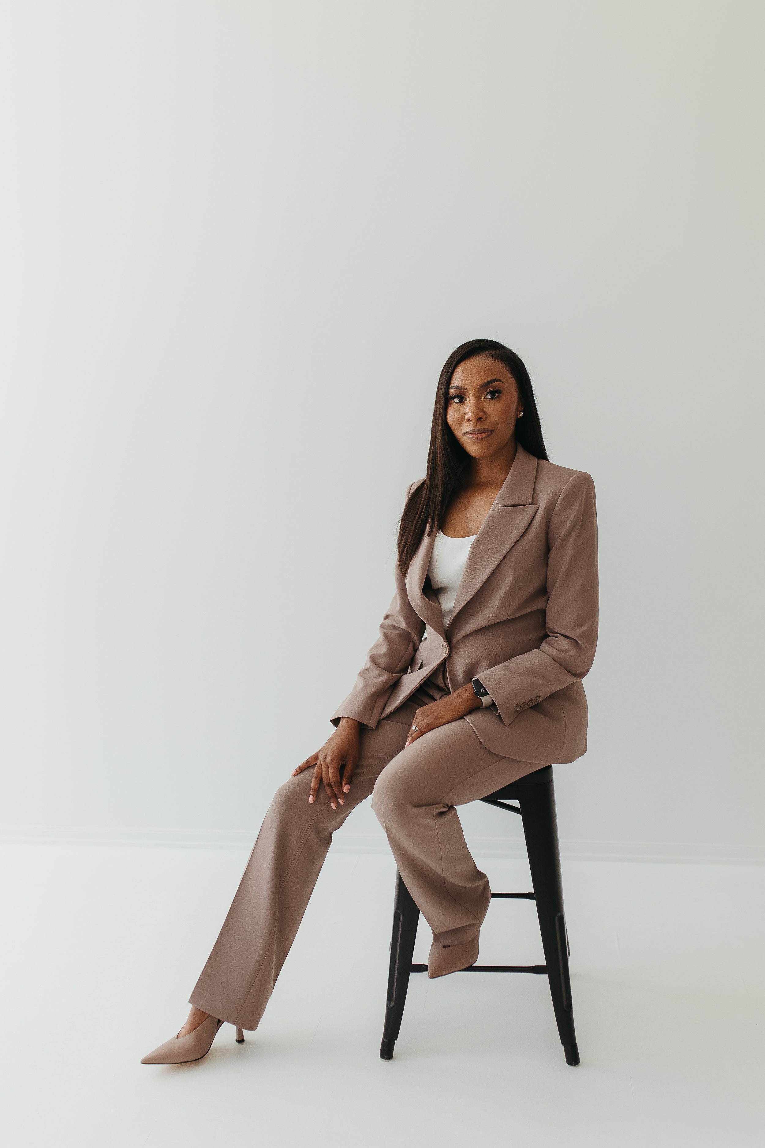 A woman in a taupe business suit sitting on a black stool in front of a plain white wall.