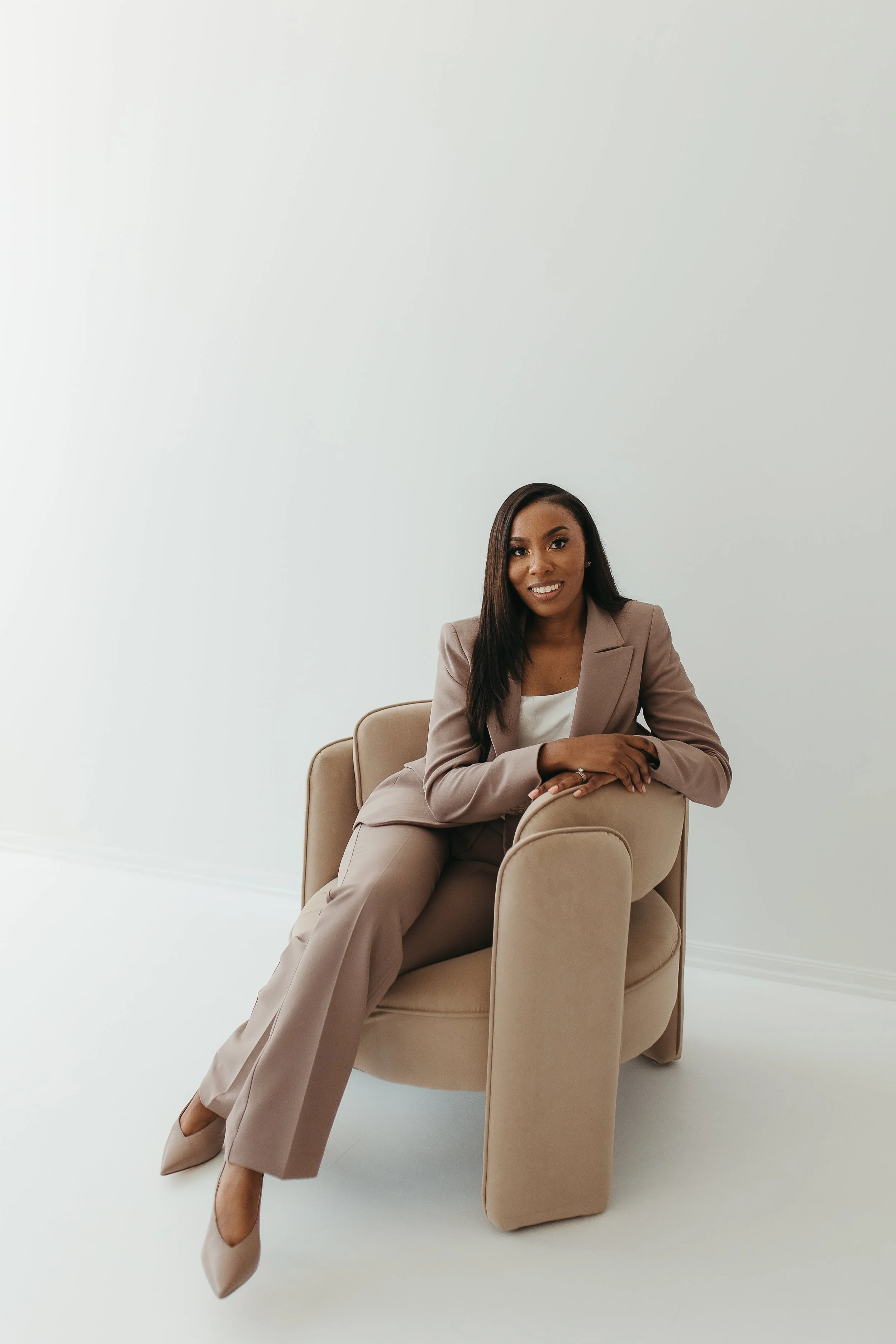 A woman in a beige pantsuit sitting on a matching beige armchair against a plain white wall.