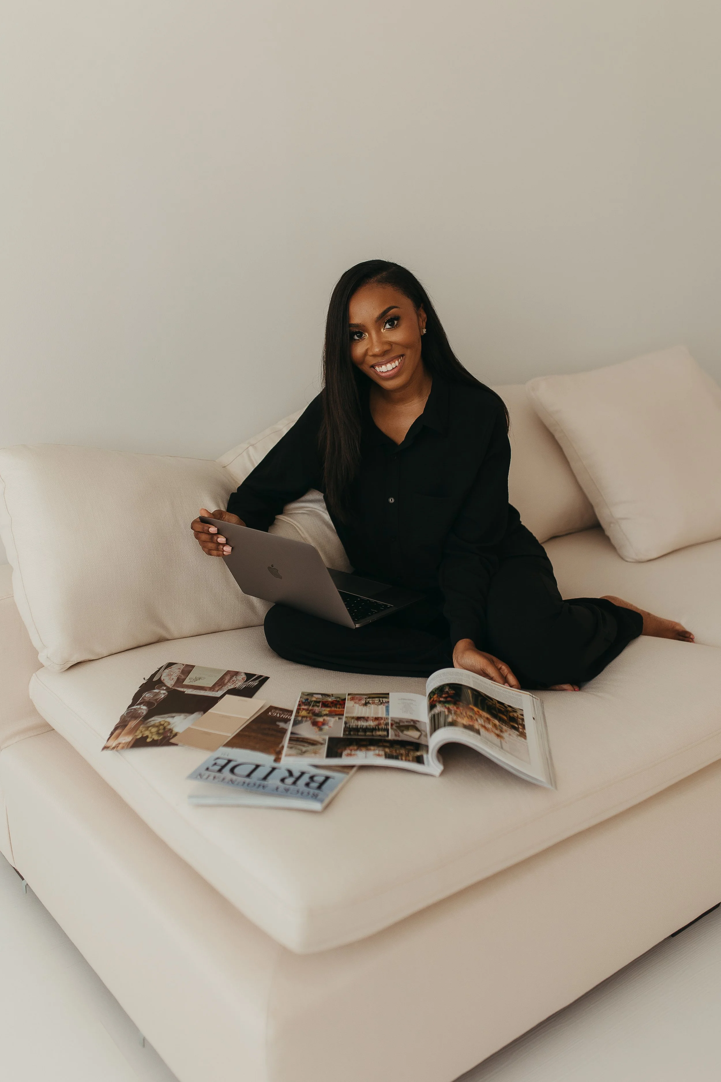 A woman sitting on a white sofa holding a laptop, with open magazines and a book spread out on the sofa in front of her, smiling at the camera.
