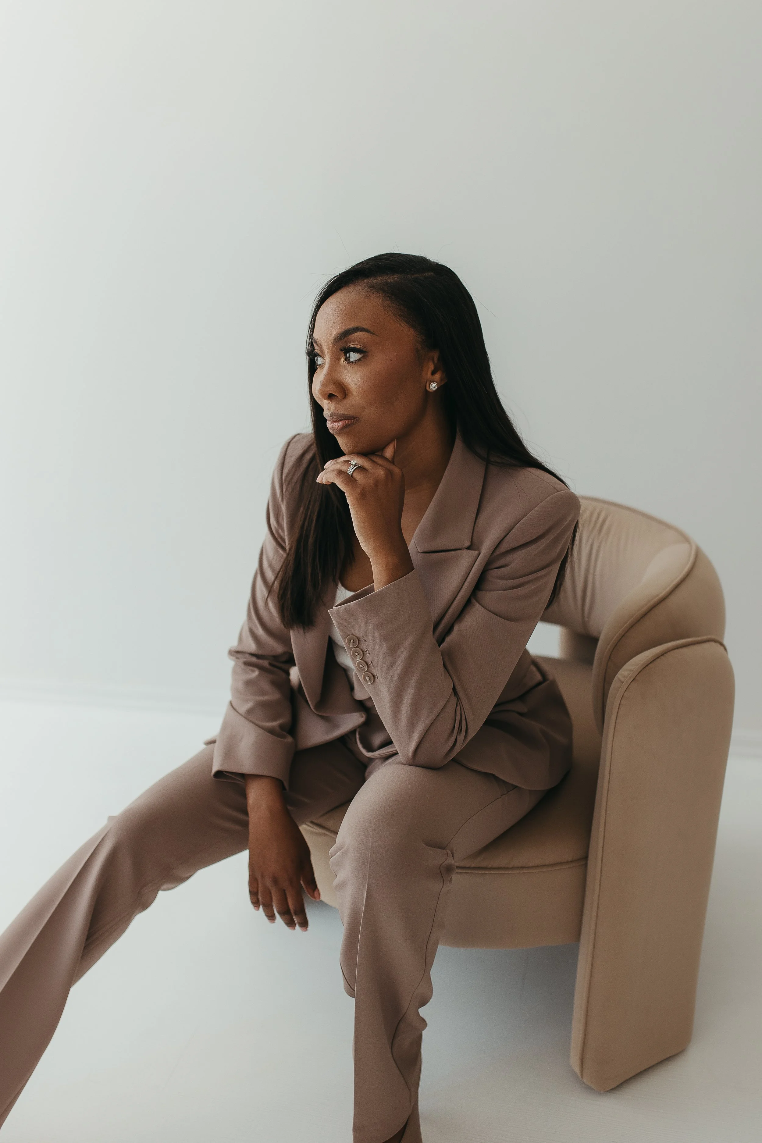A woman in a beige business suit sitting on a beige chair, with her chin resting on her hand, looking thoughtful against a plain white background.