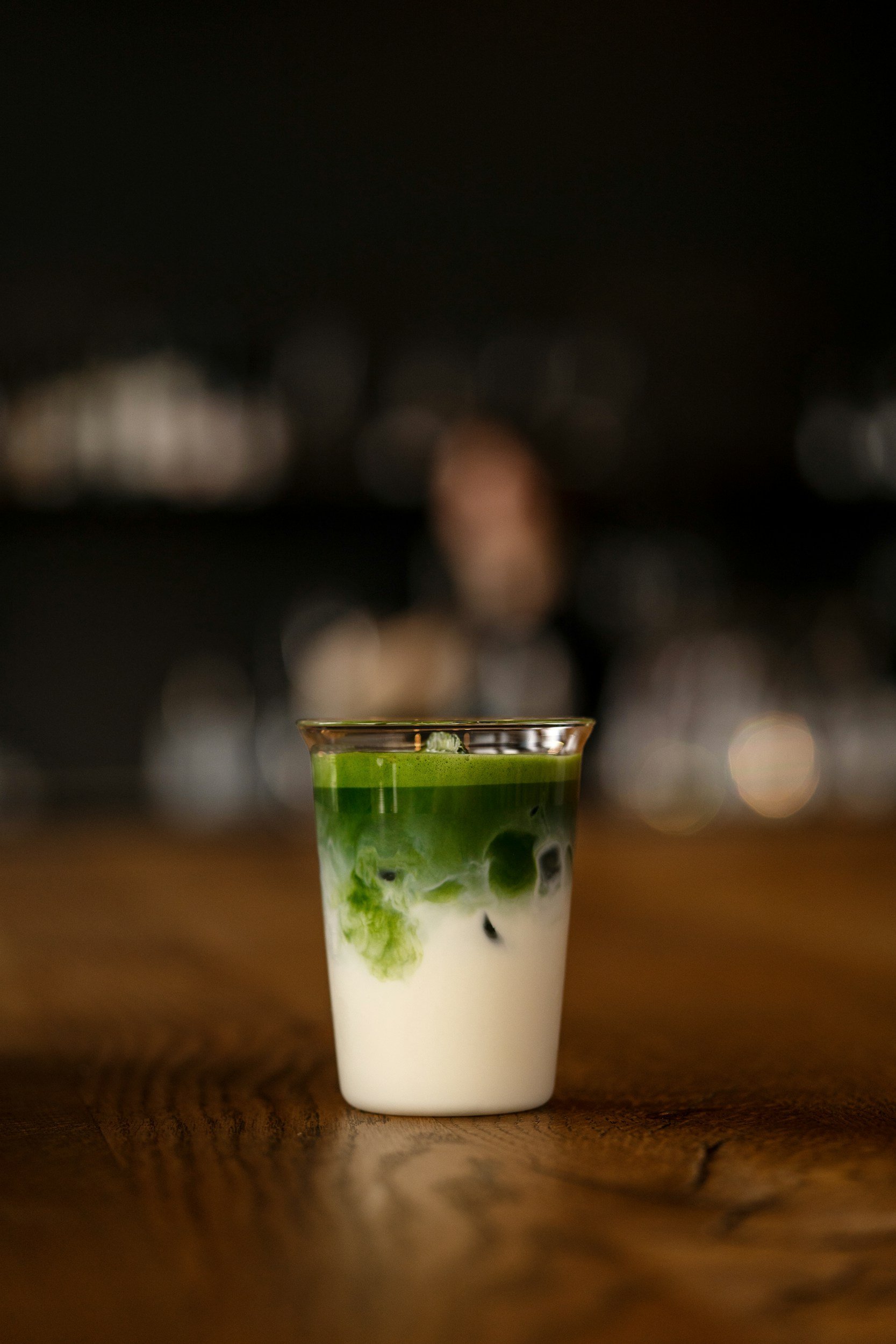 A glass filled with layered matcha, milk, and black tapioca pearls sits on a wooden table with a blurred cityscape background at night.