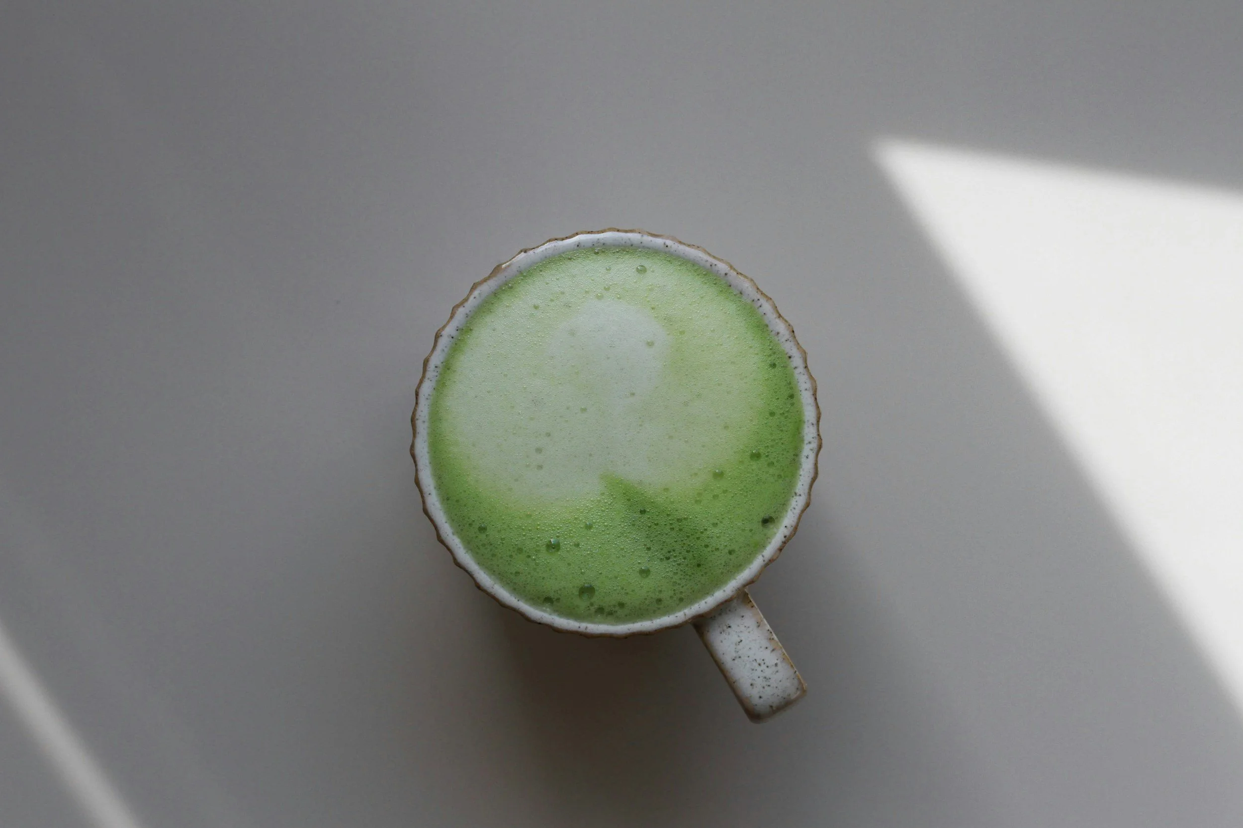 Top view of a ceramic cup filled with frothy matcha green tea on a light-colored surface, with a shadow cast on the right side.
