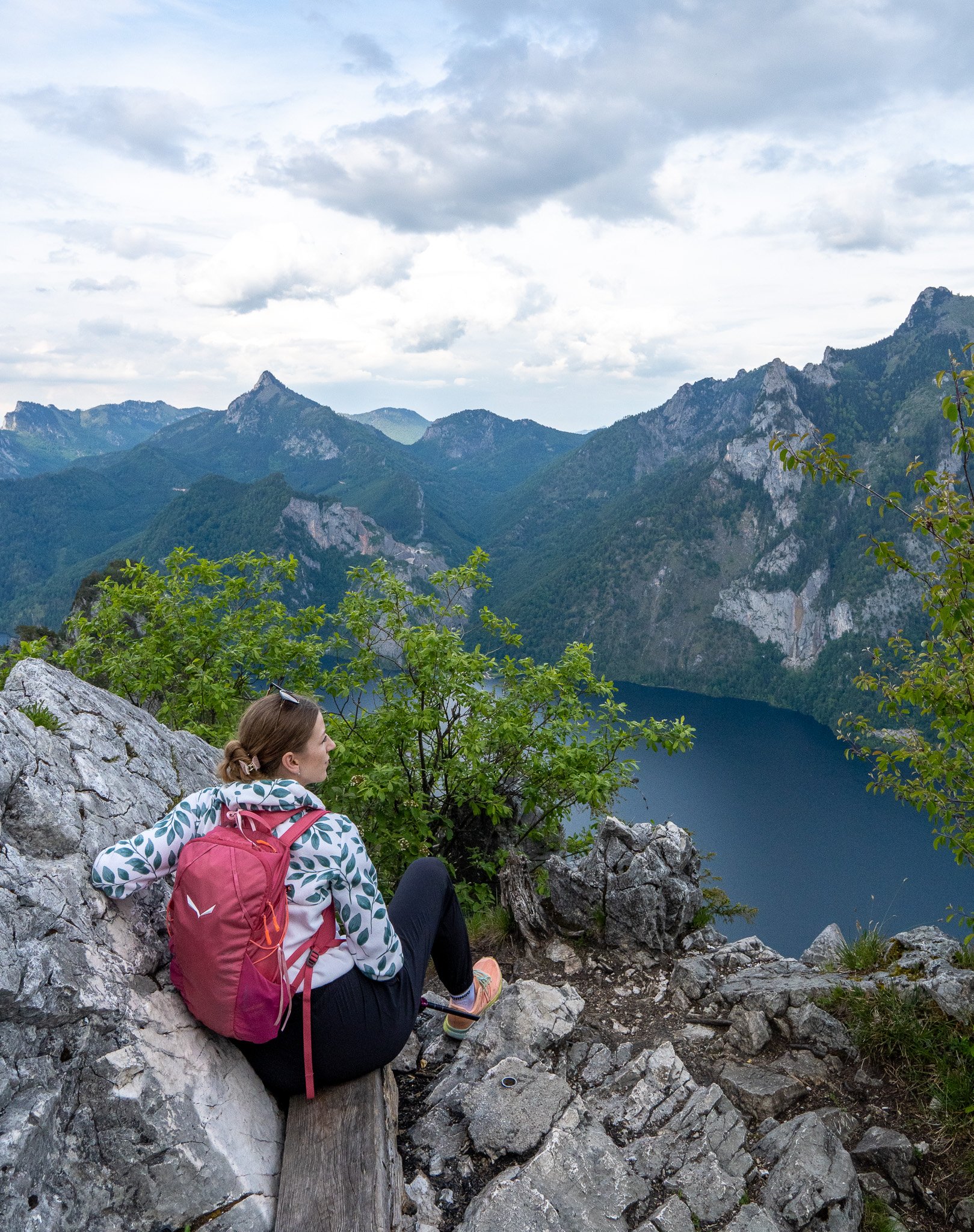 A woman with a pink backpack and leopard print jacket sitting on rocky terrain near a mountain lake, overlooking lush green mountains and partly cloudy sky.