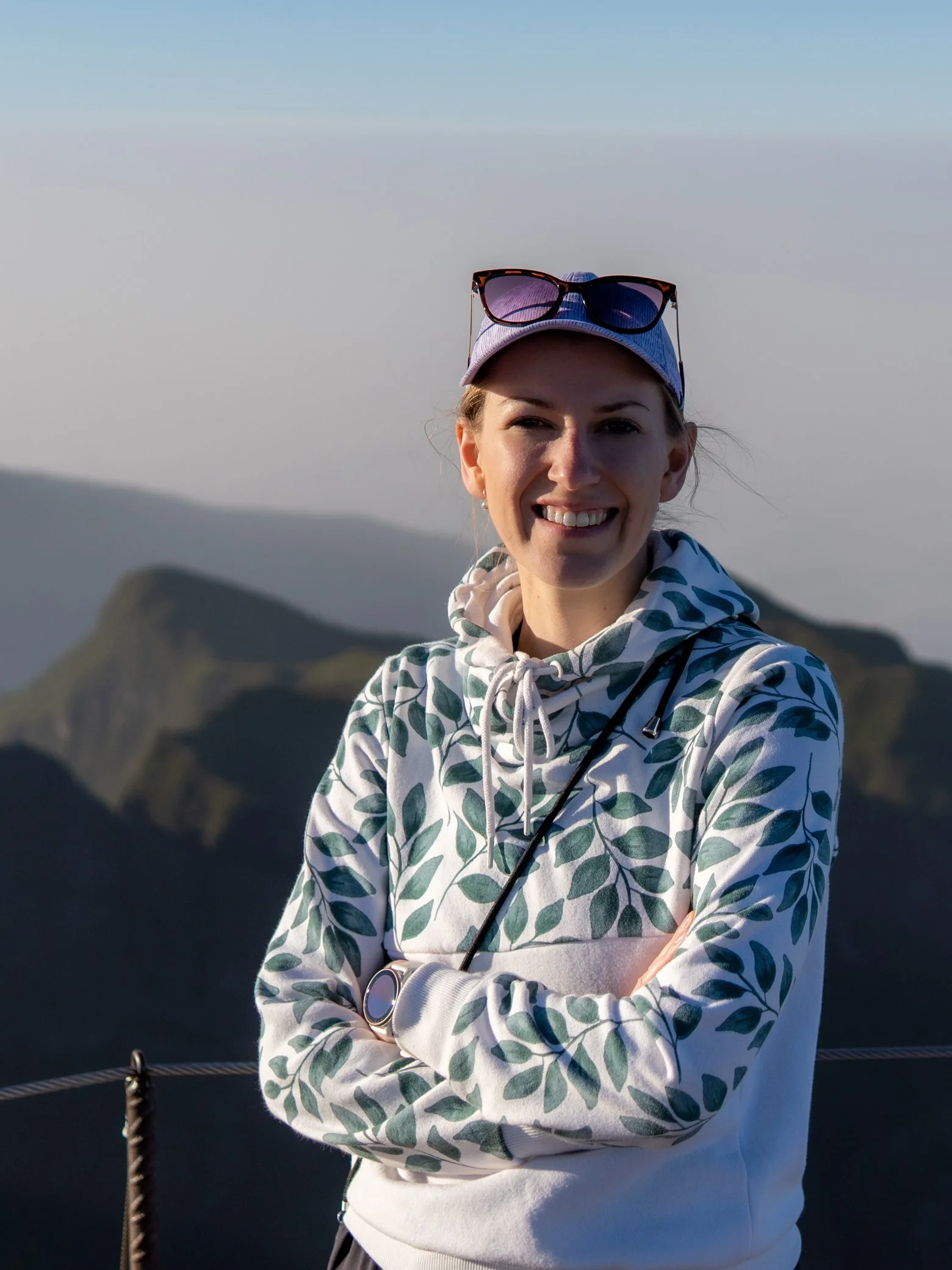 A young woman in a white hoodie with blue leaf patterns, a white cap with sunglasses on top, stands on a mountain with a blurred mountain landscape in the background.