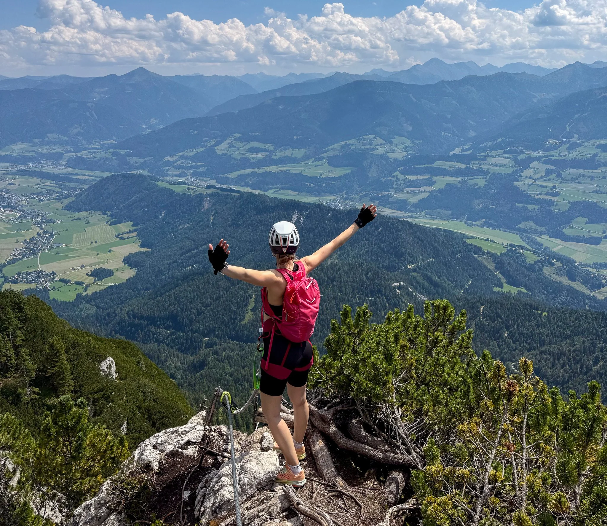A person wearing a helmet and pink backpack hiking on a rocky trail on a mountain, raising their arms, with a view of expansive mountains, valleys, and clouds in the background.