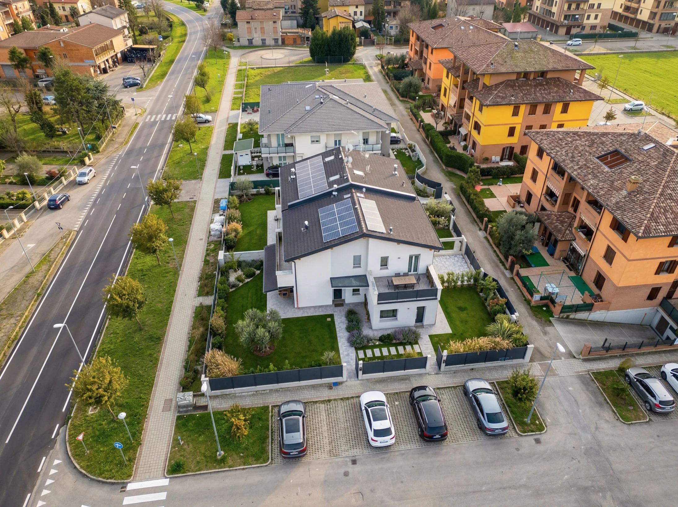 Vista aerea di un complesso residenziale con case di diversi stili e colori, un'area verde centrale con alberi, un parcheggio con auto e una strada che la attraversa. Modena cantiere concluso