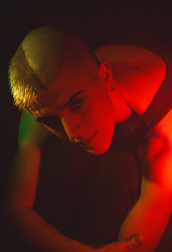 A person with short, bleached hair in a dark setting illuminated by red and green lighting, looking at the camera with intense expression, wearing a sleeveless top and earrings.
