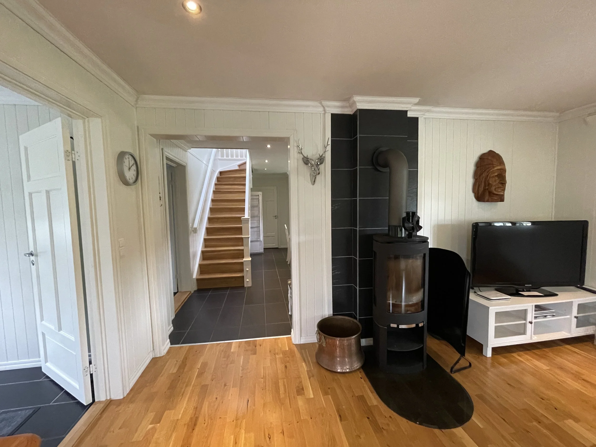 Blick in ein Wohnzimmer mit Kamin und Treppe im Hintergrund, weiße Holzvertäfelung, Wanddekorationen, Fernseher, Möbel und Holzboden.