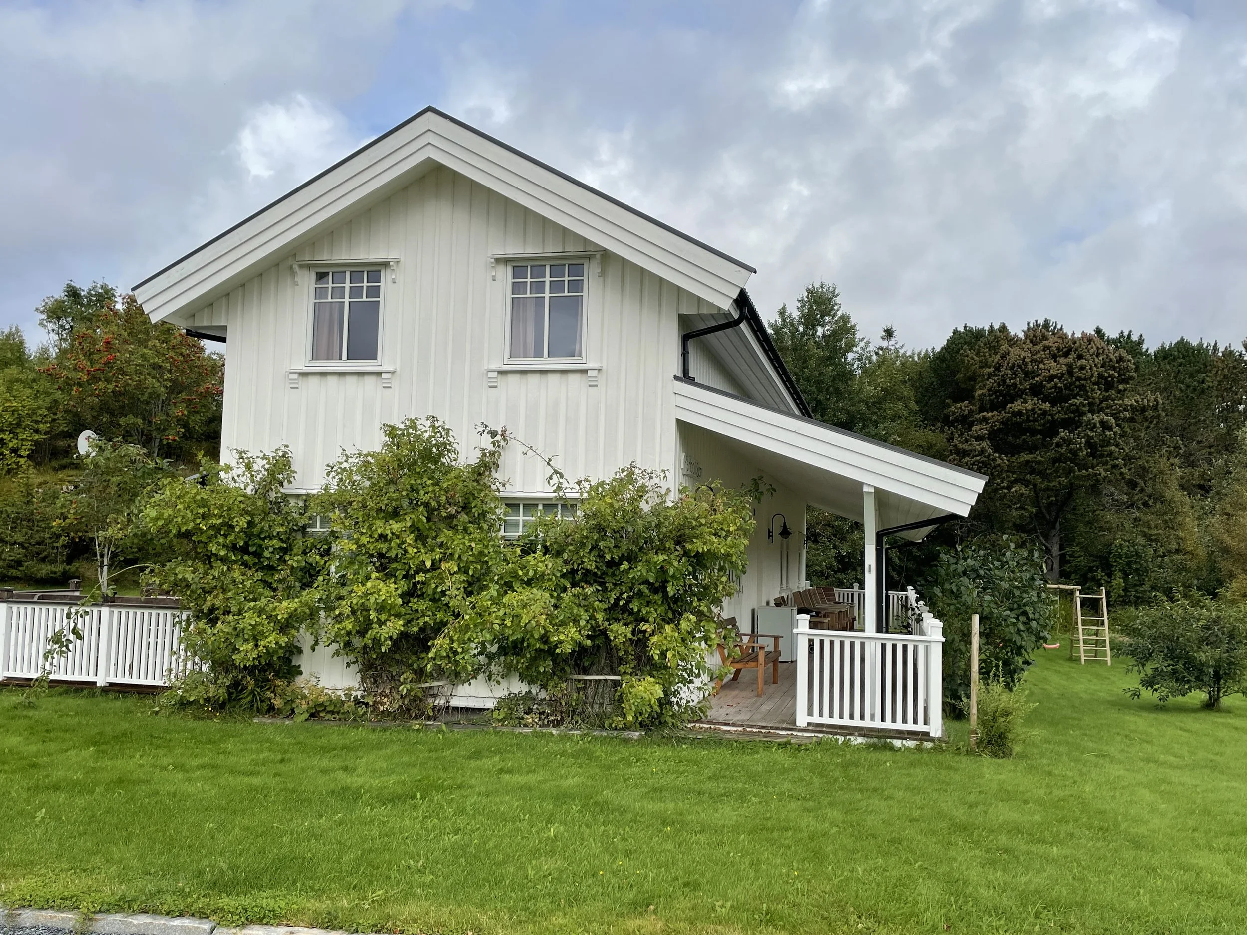 Ein weißes, zweistöckiges Haus mit großen Fenstern, umgeben von einem Garten mit Rasen, Büschen und Bäumen. Es gibt eine Terrasse mit Gartenmöbeln und eine aufgestellte Schaukel im Garten.