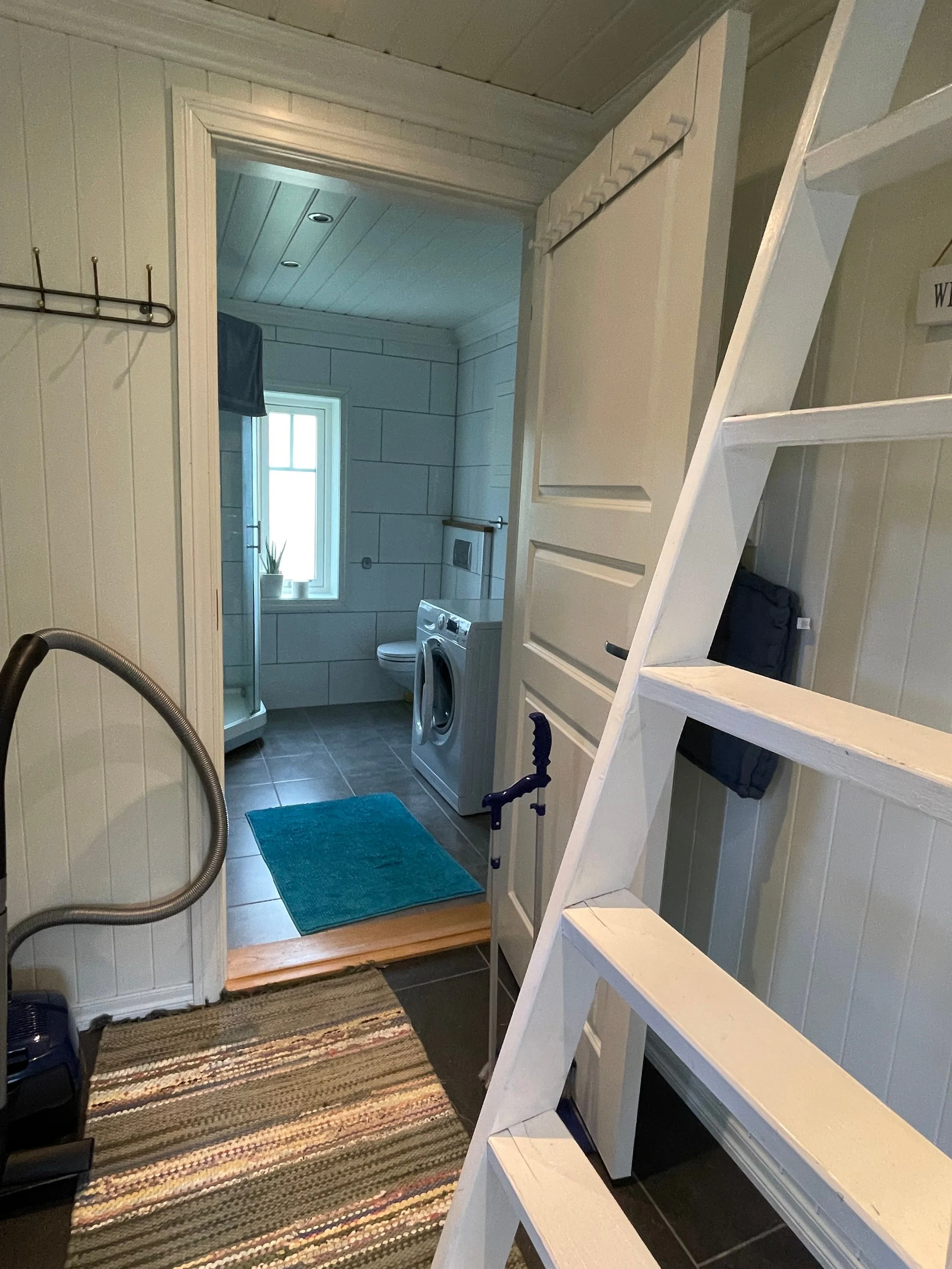 Blick in ein Badezimmer mit Waschmaschine, Dusche, kleinen Fenster und weißen Wandpaneelen, vor einem Treppenabsatz mit buntem Teppich.