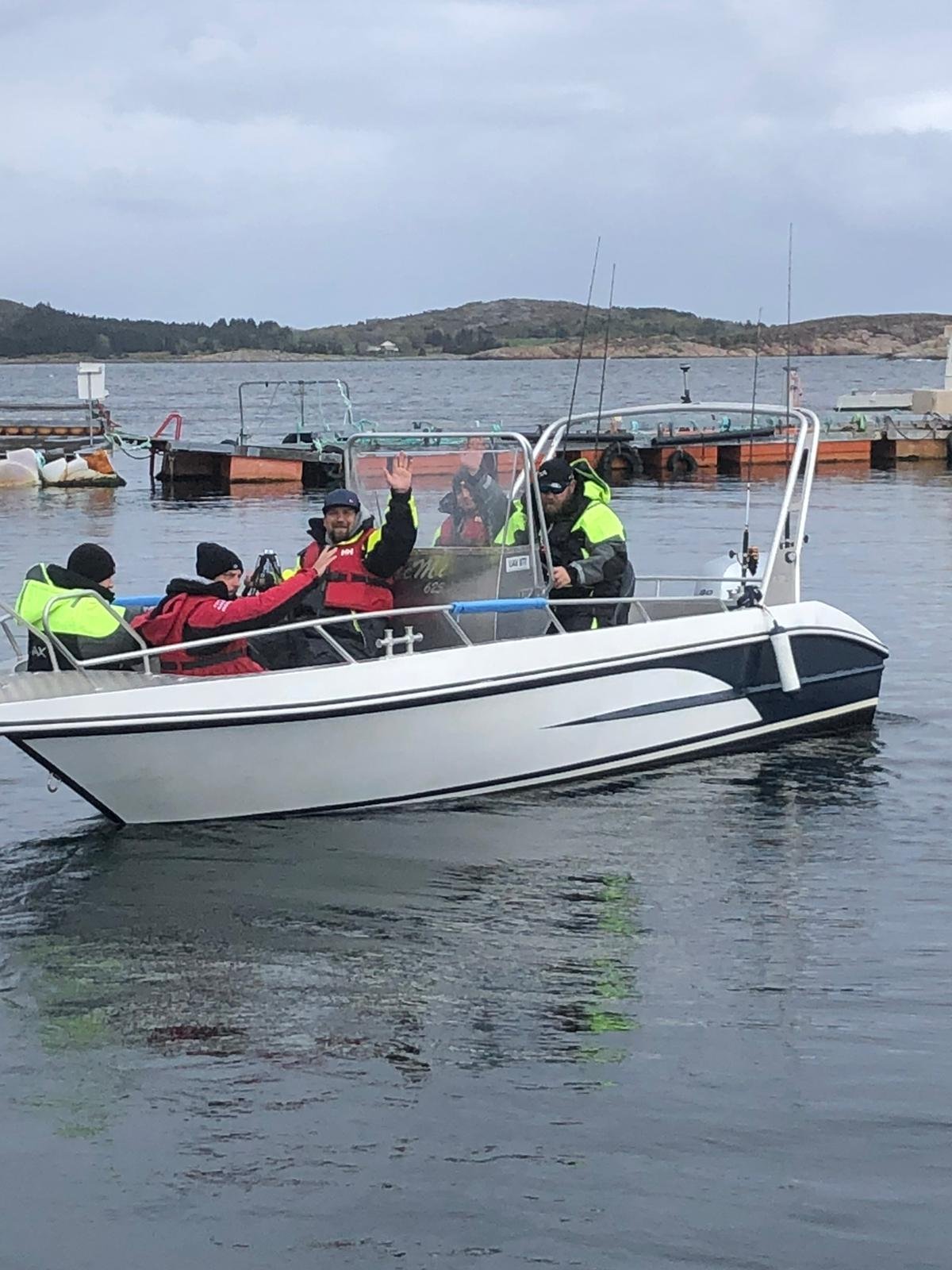 Menschen auf einem Boot mit gelber und roter Sicherheitsweste auf dem Wasser, im Hintergrund Felsen und Boote am Steg.