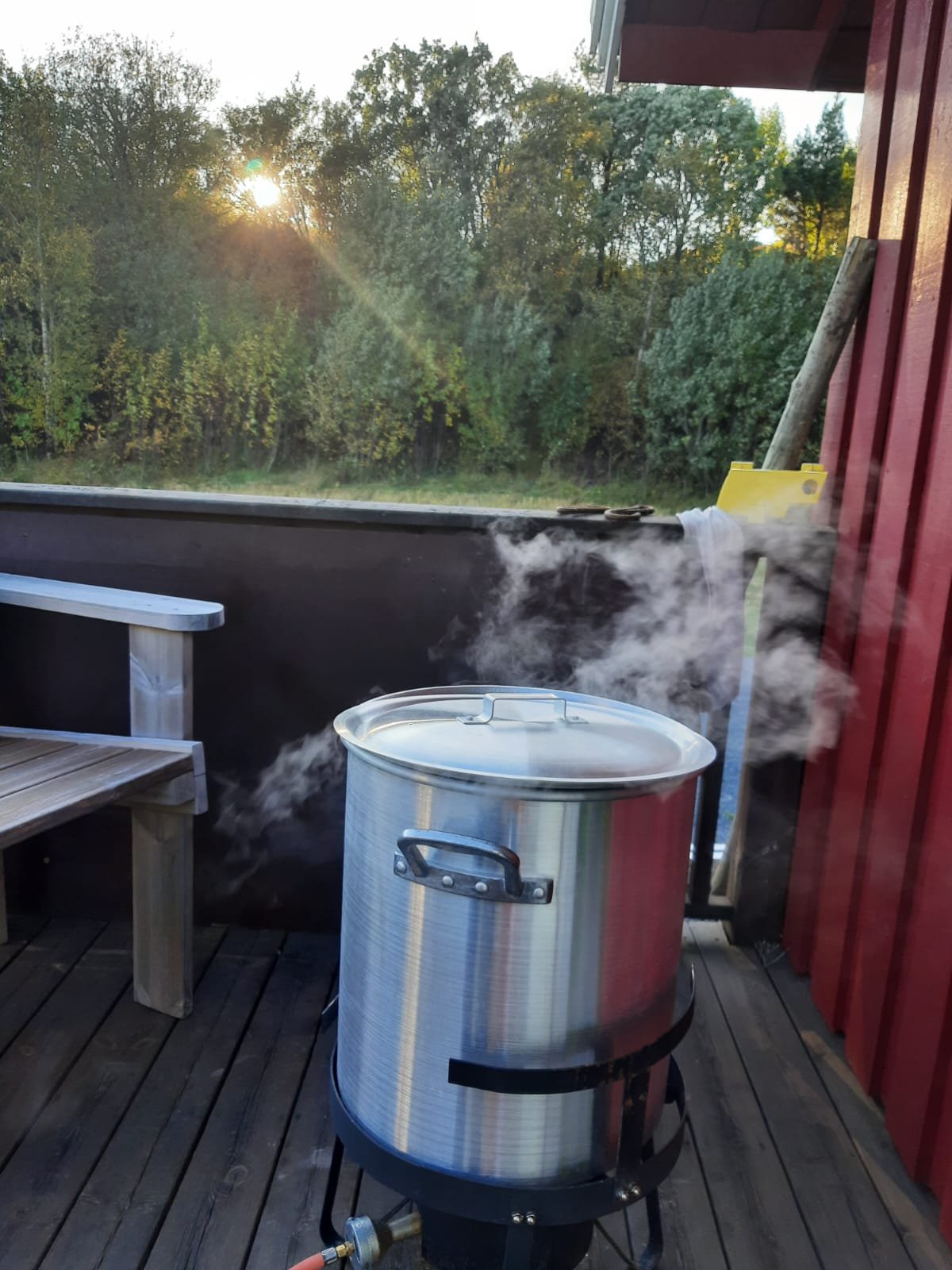 Ein großer silberner Topf auf einem Heizofen auf einem Holzdeck, mit sichtendem Dampf, im Freien bei Sonnenuntergang, im Hintergrund Bäume.