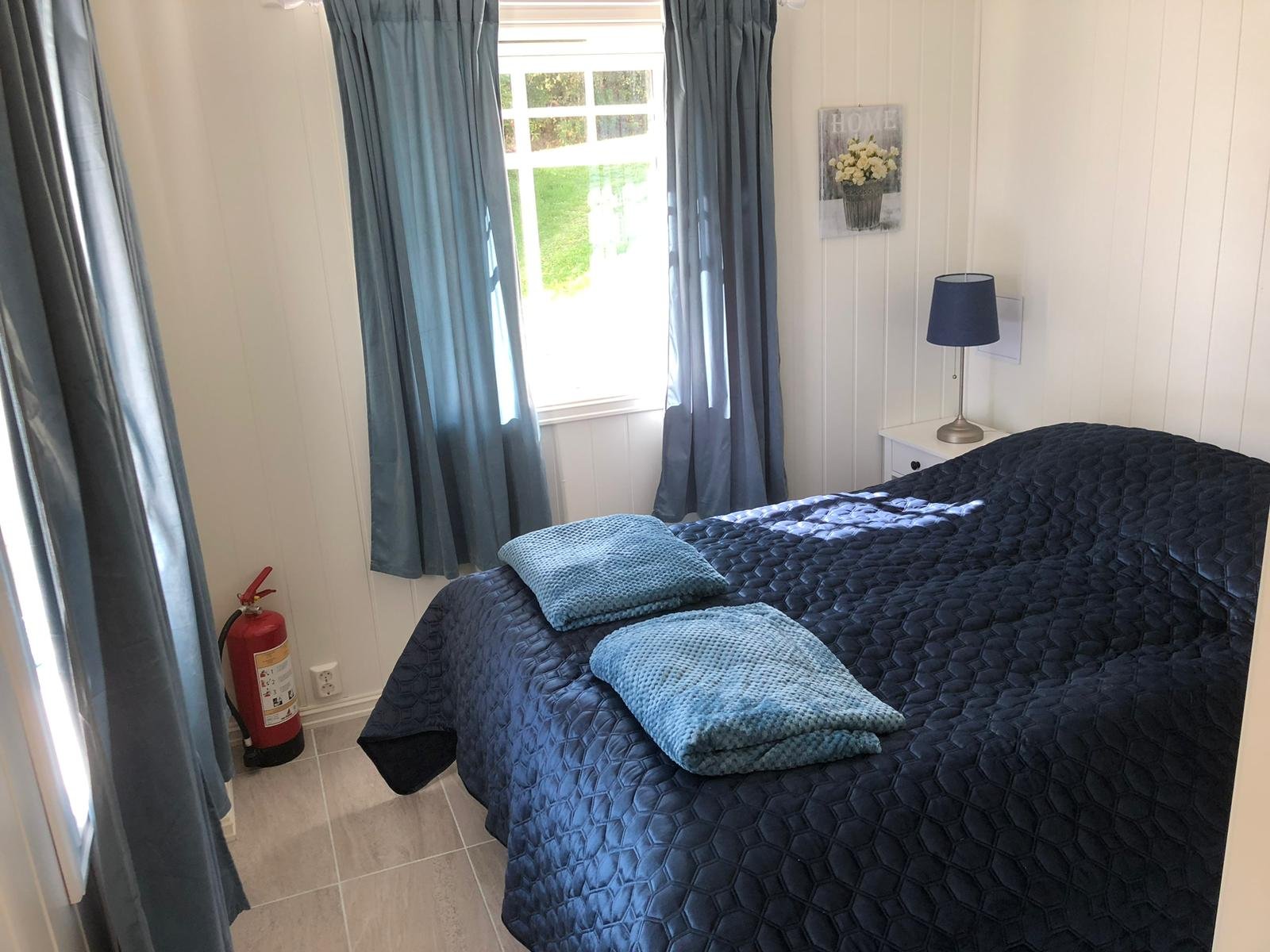 Ein Schlafzimmer mit schwarzem Bett, blauen Kissen, Nachttischlampe, Fenster mit blauen Vorhängen, Wanddeko mit Blumen, Feuerlöscher im Eck, weiße Wände.