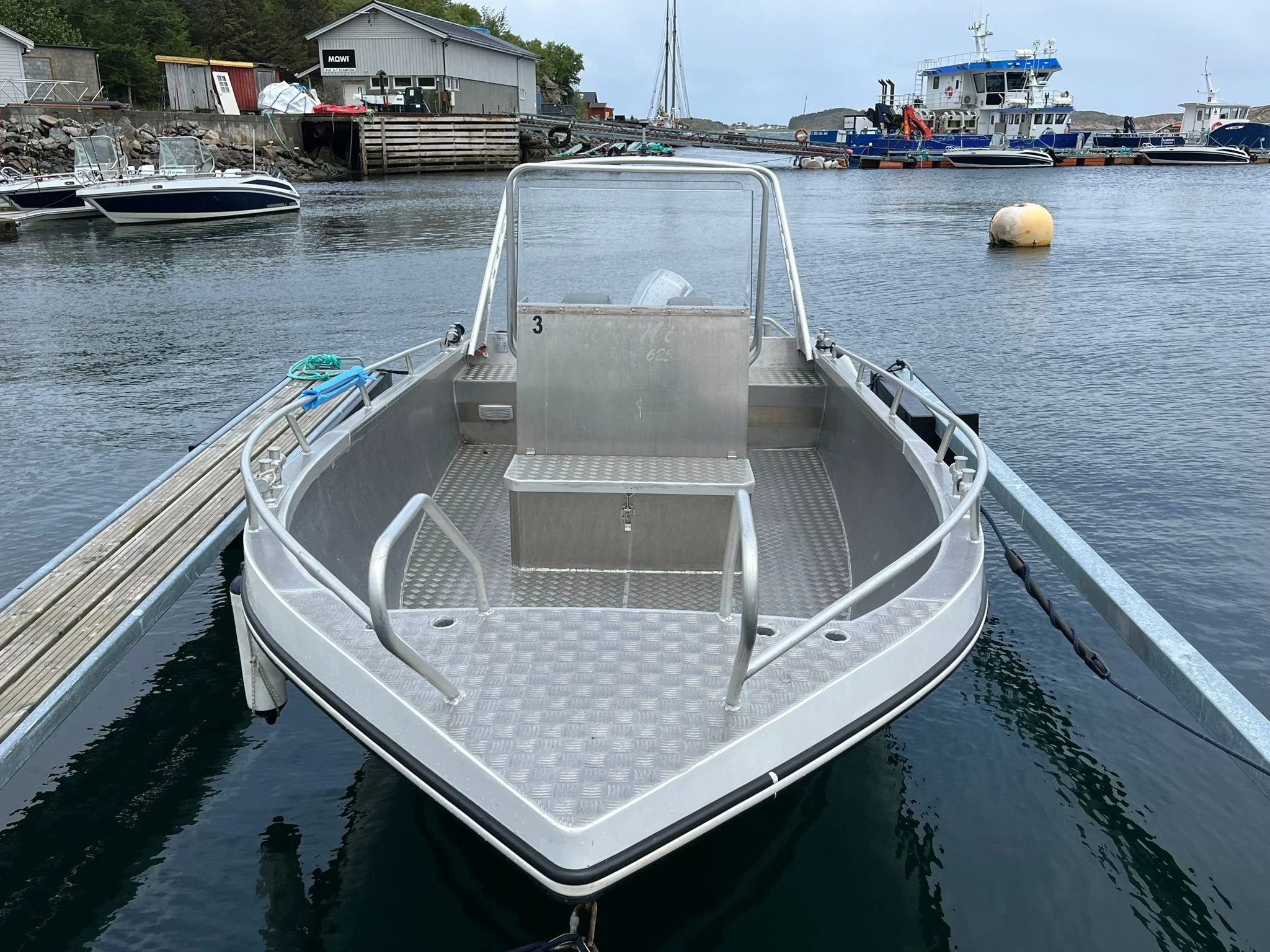 Leeres Aluminium-Boot im Hafen, umgeben von Wasser und weiteren Booten, mit einem Gebäude im Hintergrund.