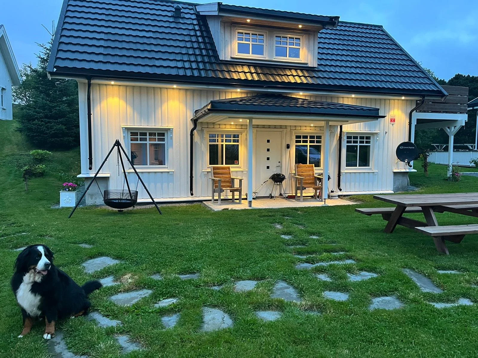 Ein weißes, zweistöckiges Haus mit schwarzem Ziegeldach, das bei Dämmerung mit Außenbeleuchtung erleuchtet ist. Im Garten sind eine Schaukel, ein Grill, ein Picknicktisch und ein Border Collie Hund sichtbar.