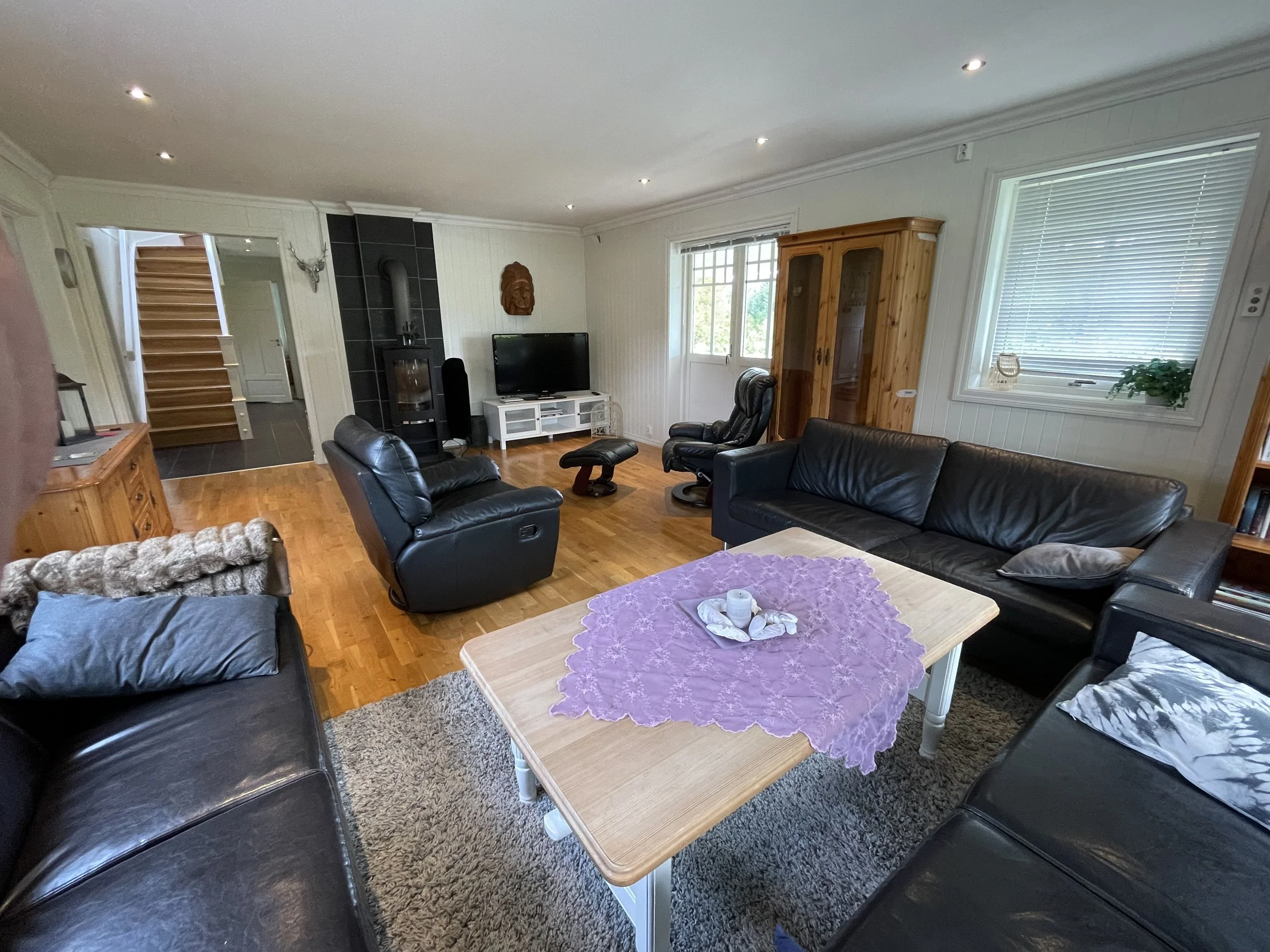 Ein Wohnzimmer mit schwarzem Leder-Couch, einem Fernseher auf einem weißen TV-Schrank, einem Holzzustand und mehreren Fenstern. Es gibt einen stilvollen Kaminkamin und dekorative Elemente an den Wänden.