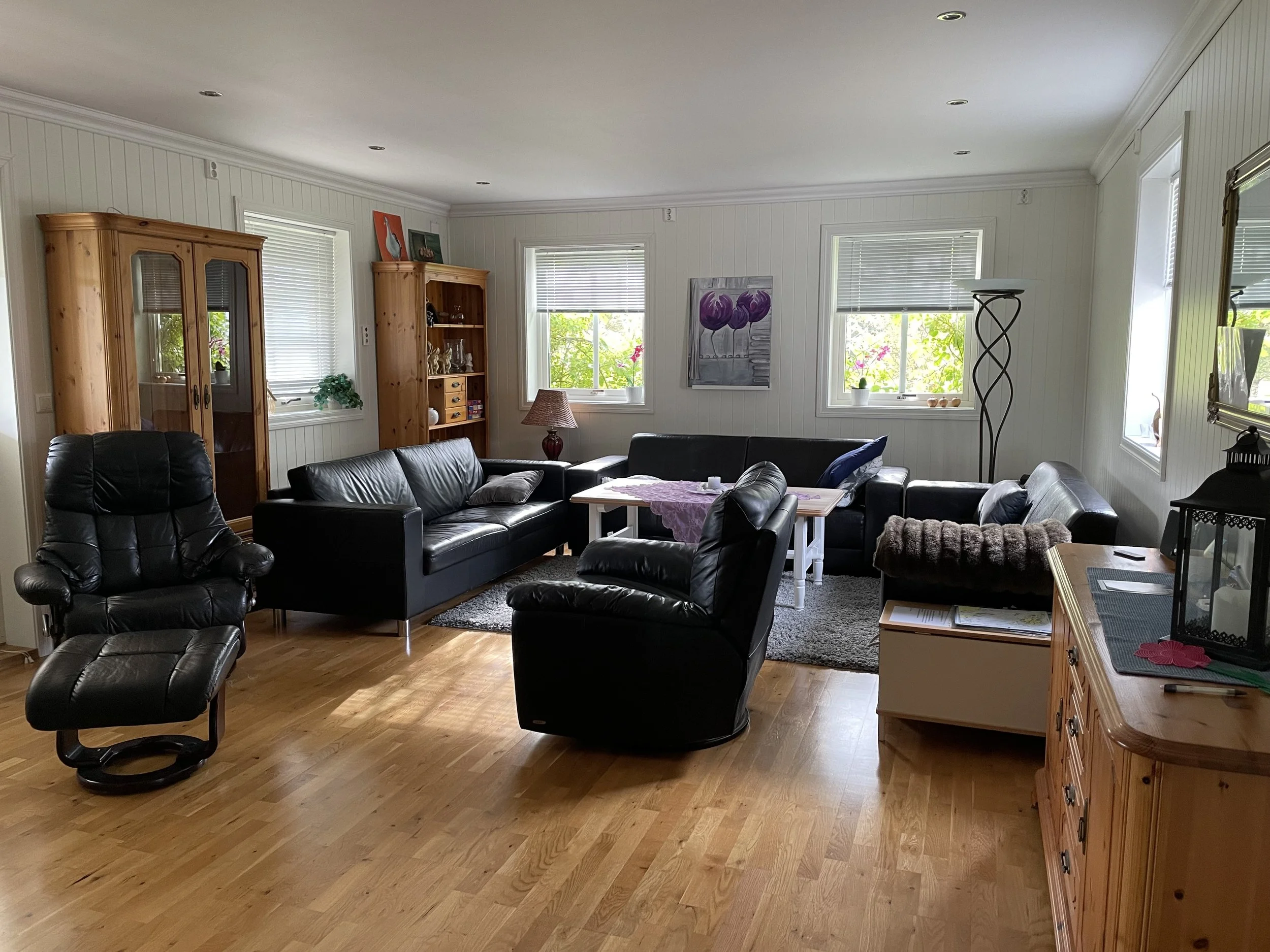 Ein Wohnzimmer mit Holzboden, schwarzen Ledersofas, einem Couchtisch, einer Holzvitrine und einer Bücherregal, Fenster mit Rollos, Wandkunst und Dekorationsgegenständen.