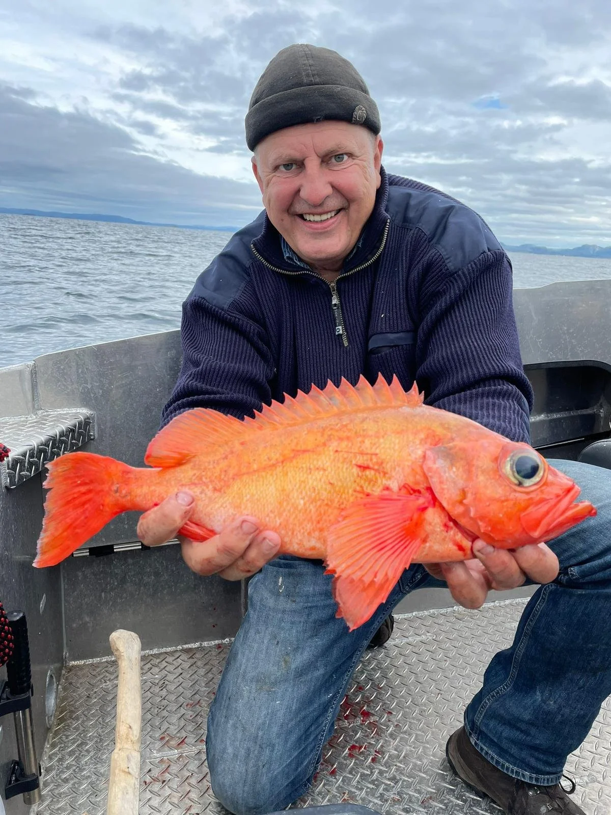 Mann auf einem Boot, hält einen großen orange-roten Fisch vor sich, im Hintergrund Wasser und Himmel mit Wolken.