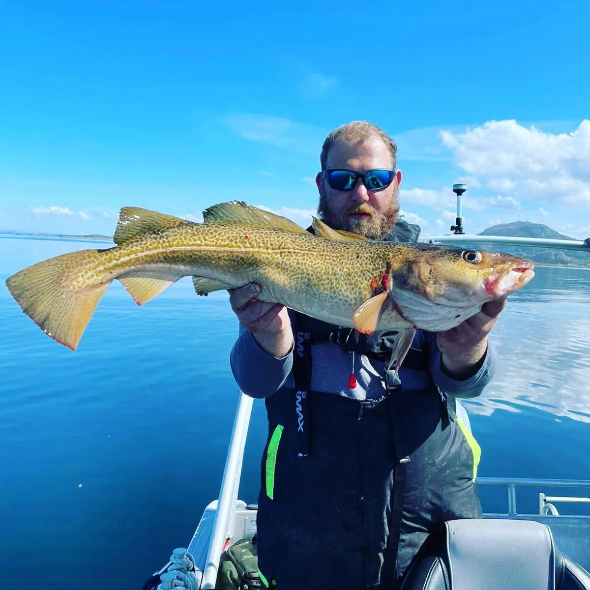 Mann mit Sonnenbrille hält einen großen Fisch auf einem Boot im Wasser, Himmel und Insel im Hintergrund.