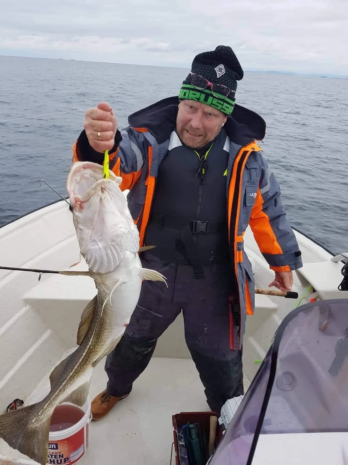 Mann auf einem Boot hält eine große gefangene Fisch mit einer Angelrute, trägt wetterfeste Kleidung und eine Mütze, im Hintergrund das offene Meer bei bewölktem Himmel.
