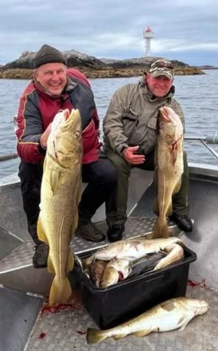 Zwei Männer auf einem Boot mit mehreren Fischern. Beide halten große gefangene Fische. Es liegt auch ein Container mit weiteren Fischen auf dem Boot, im Hintergrund ein Leuchtturm an einer felsigen Küste.