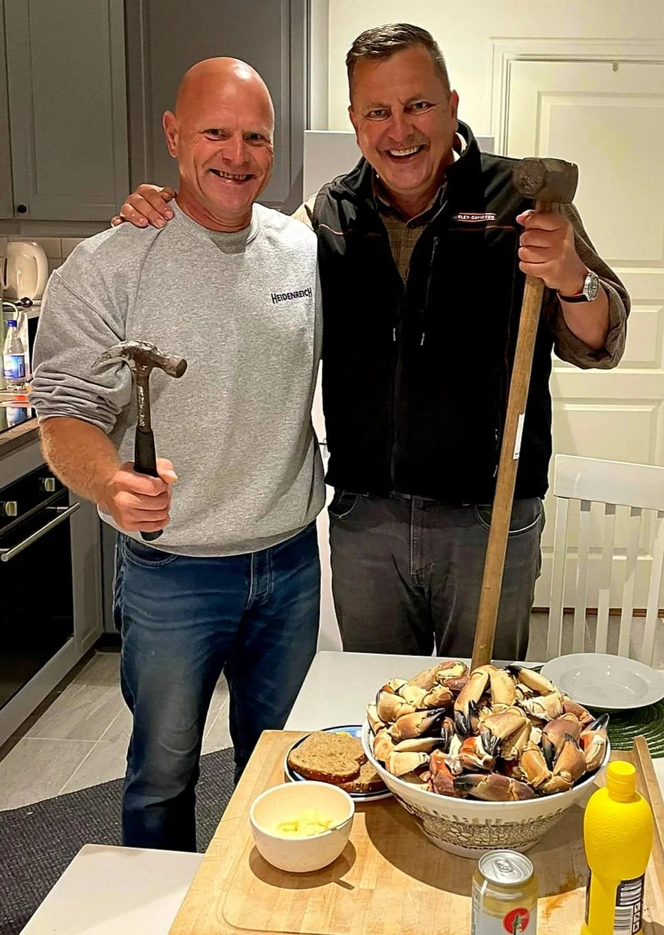 Zwei Männer stehen in einer Küche, lachen und halten Werkzeuge. Eine Schüssel mit Muscheln und ein Teller mit Toast sind auf dem Tisch, es gibt auch Butter und Senf.