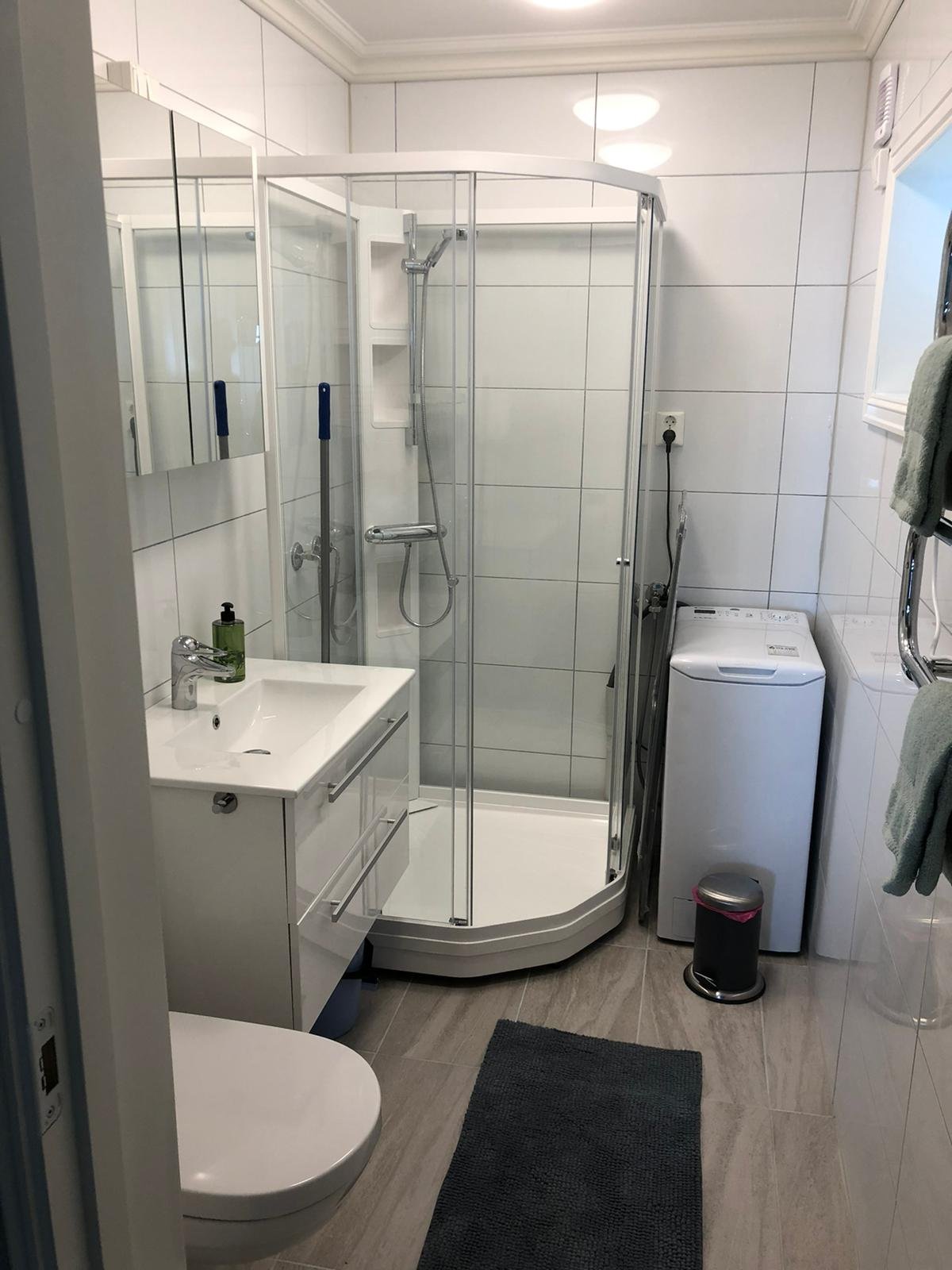 Ein kleines Badezimmer mit Dusche, Waschbecken, Waschmaschine, WC, Spiegel, Handseife und Handtüchern.