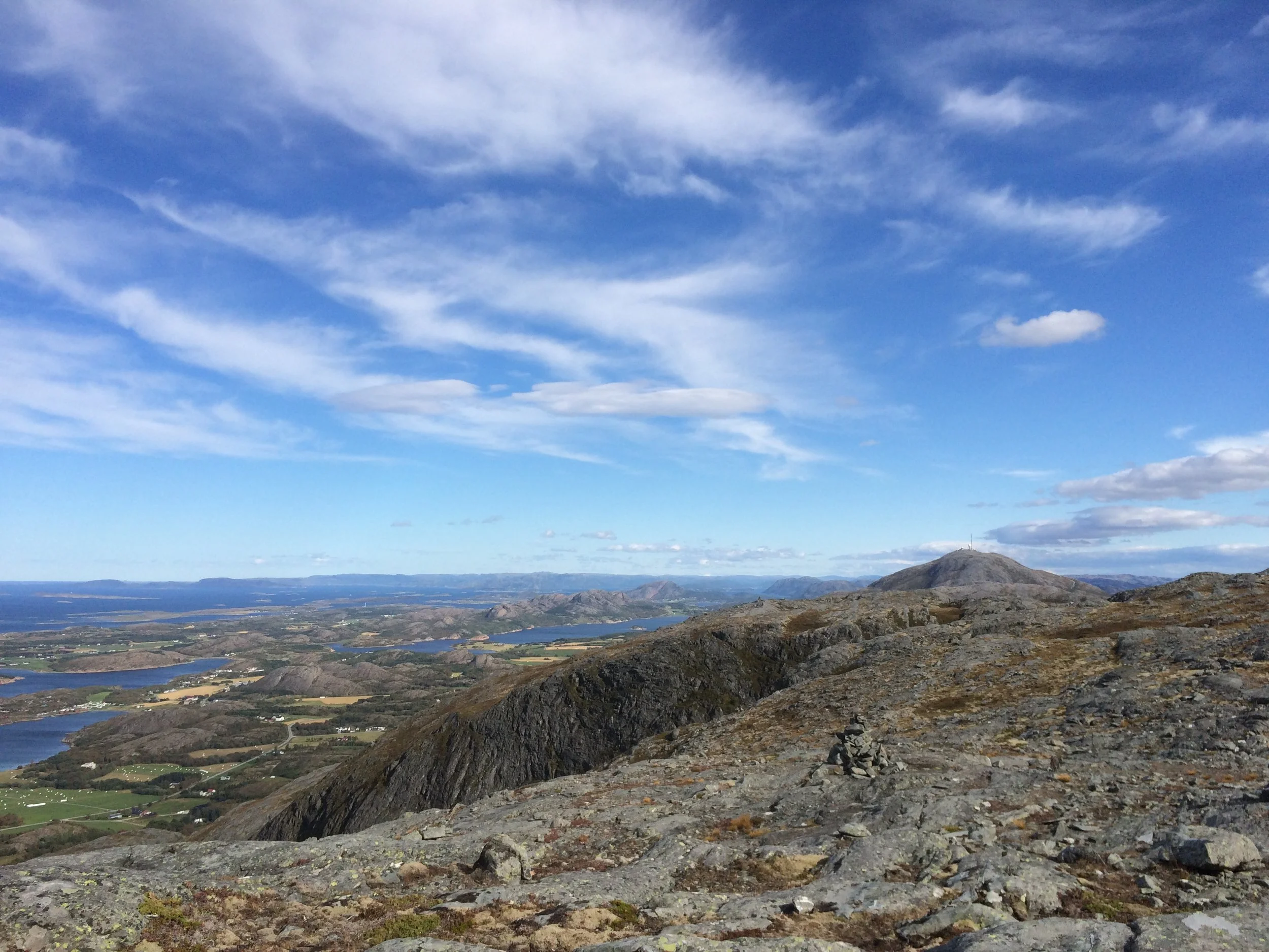 Blick von einem felsigen Berggipfel auf eine weite Landschaft mit Seen, Hügeln und Wolken im Himmel.