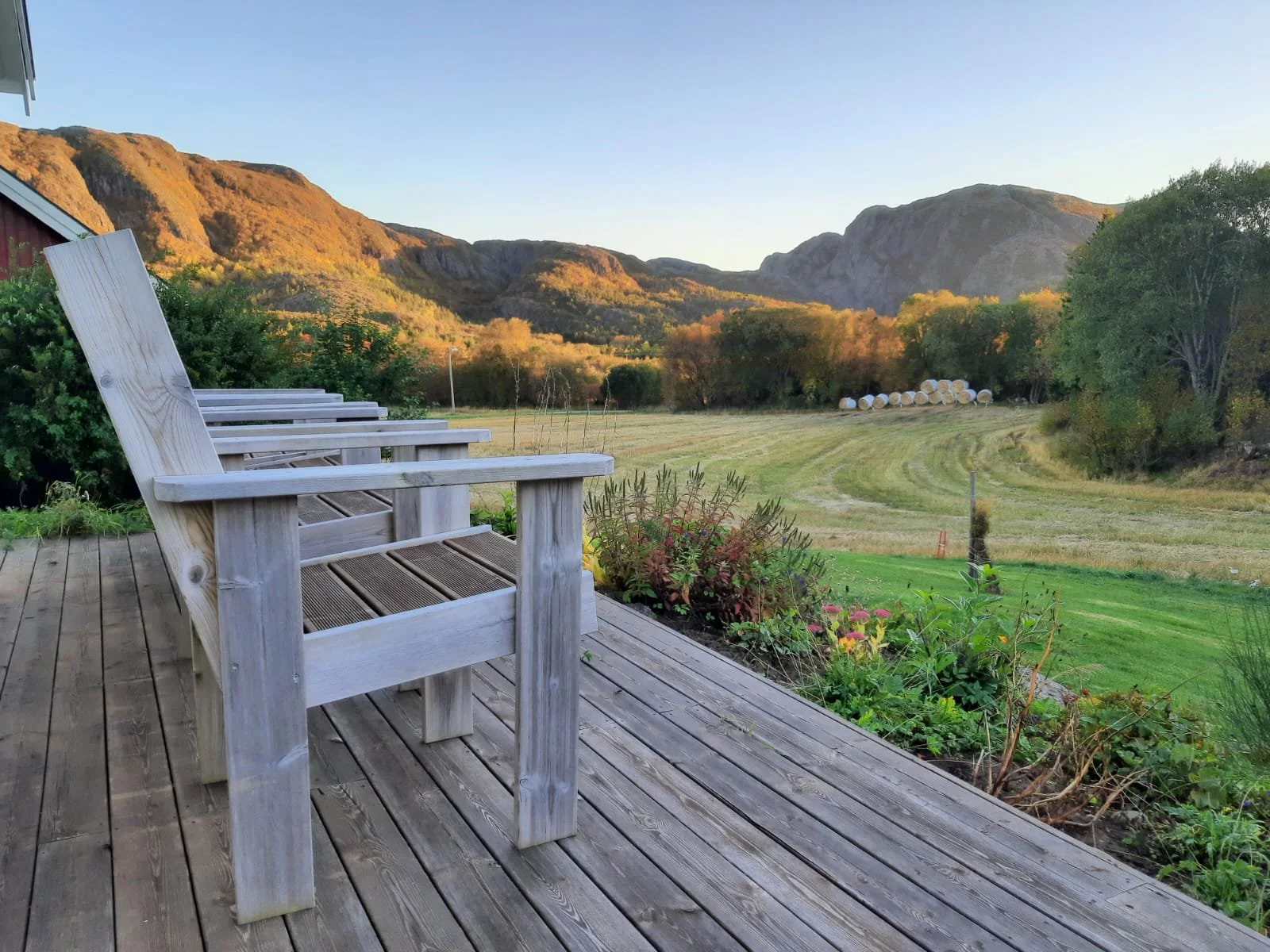 Holzterrasse mit vier leeren Holzstühlen, Blick auf eine grüne Wiese, Bäume und Berge im Sonnenuntergang.