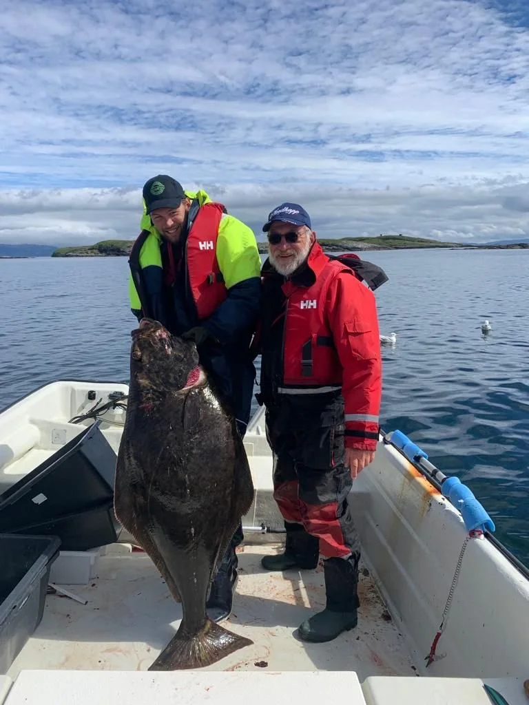 Zwei Männer auf einem Boot mit einem großen Fisch, vermutlich einem Steinbutt, vor der Küste bei schönem Wetter.
