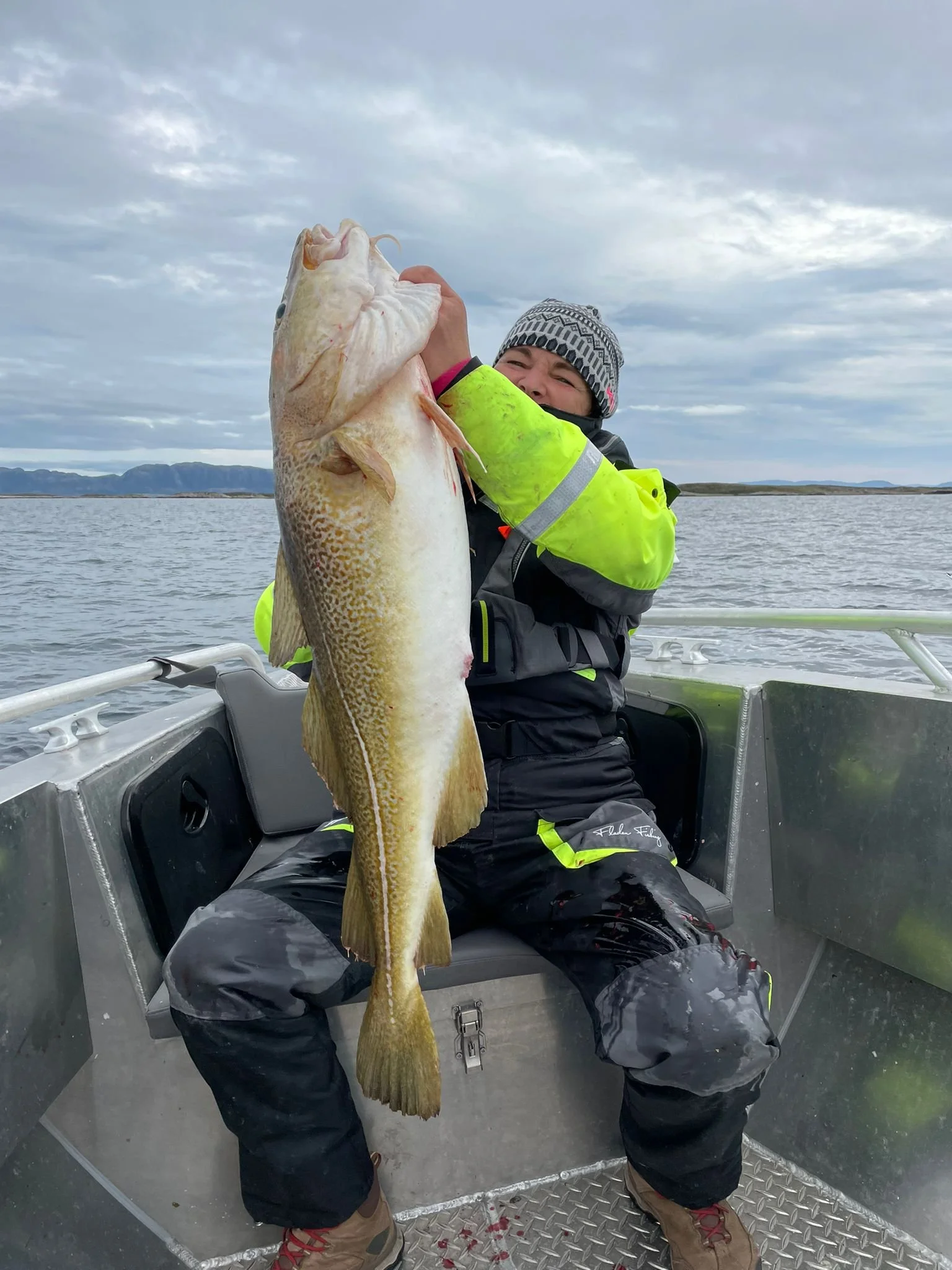 Mensch sitzt in einem Boot auf einem See und hält eine große, gefangene Fisch. Der Himmel ist bewölkt.