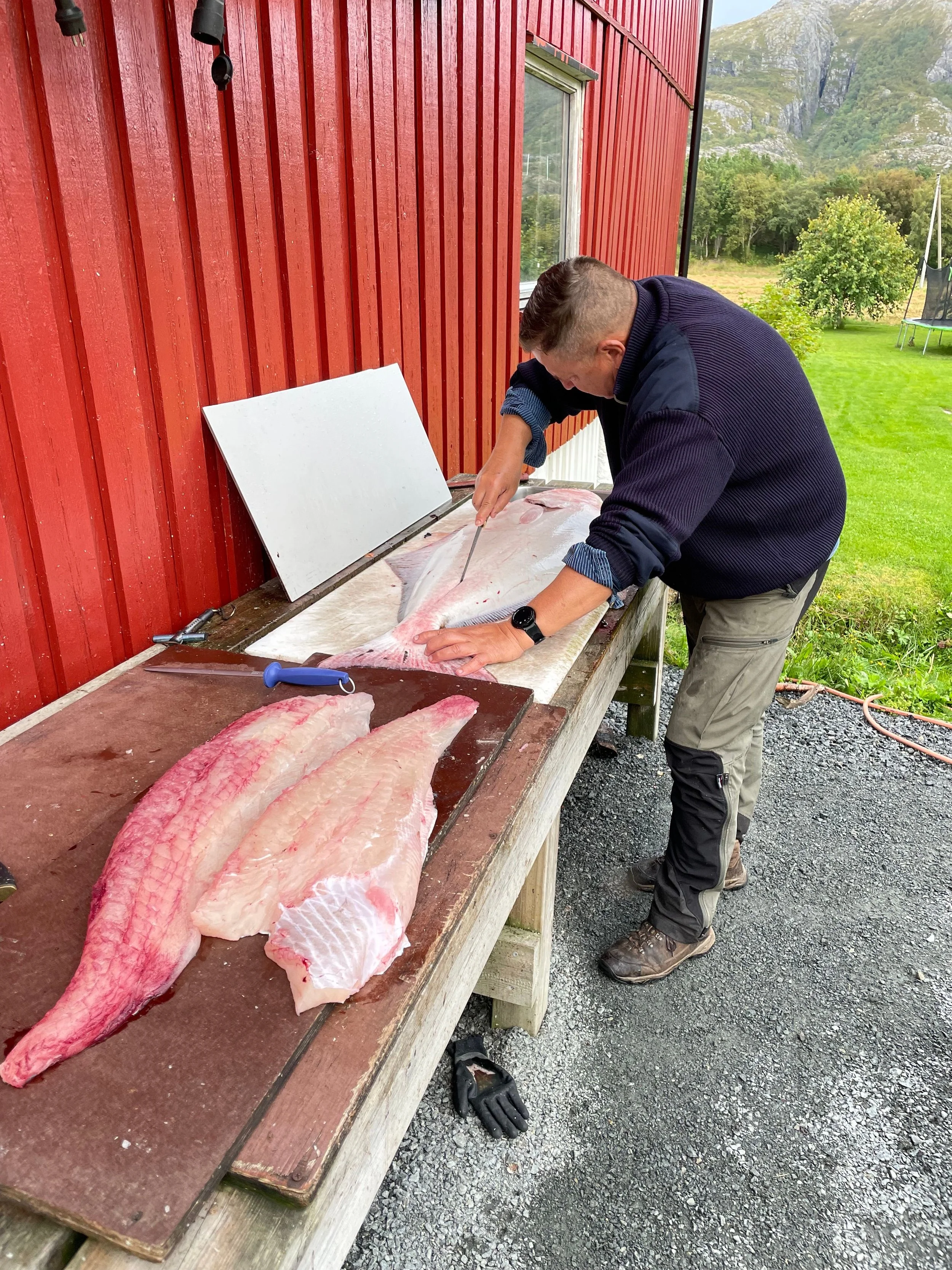 Mann schneidet Fisch auf einem Holztisch im Freien neben einer roten Holzwand