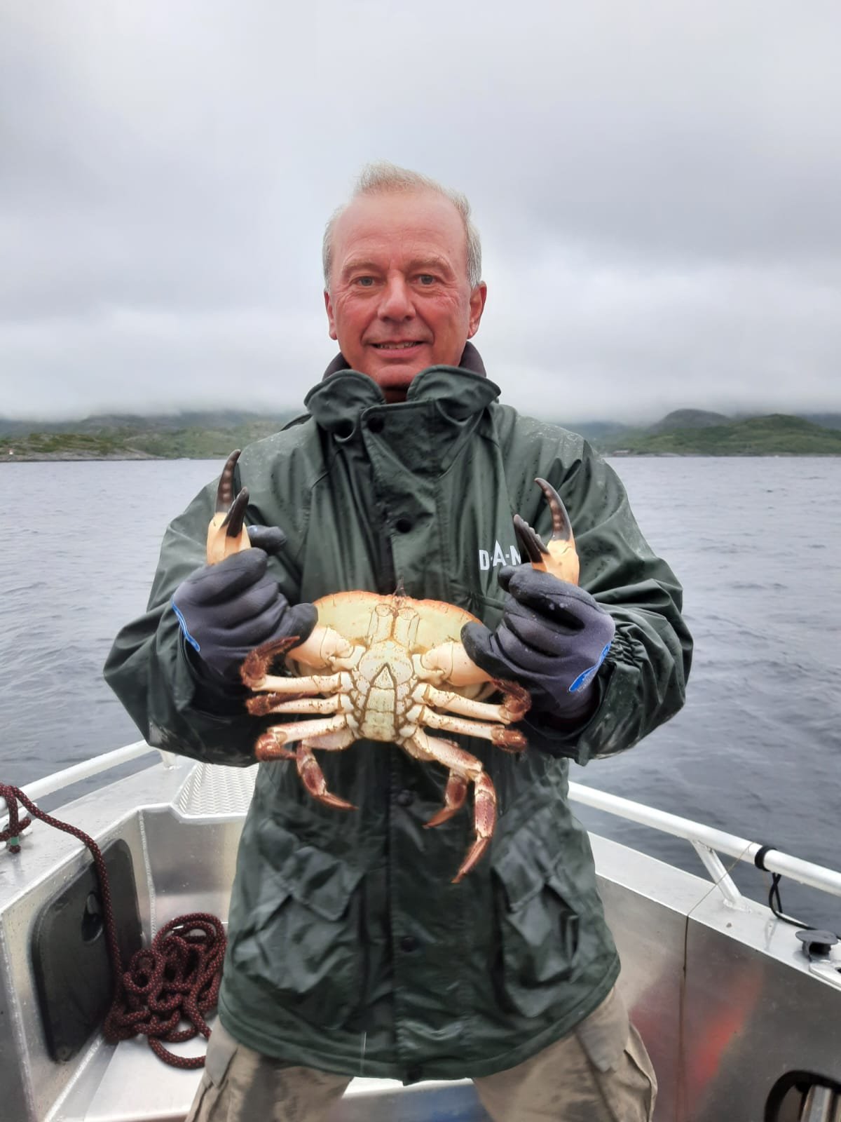 Ein Mann auf einem Boot hält eine große Krabbe in den Händen vor einem Wasserhintergrund mit bewölktem Himmel.