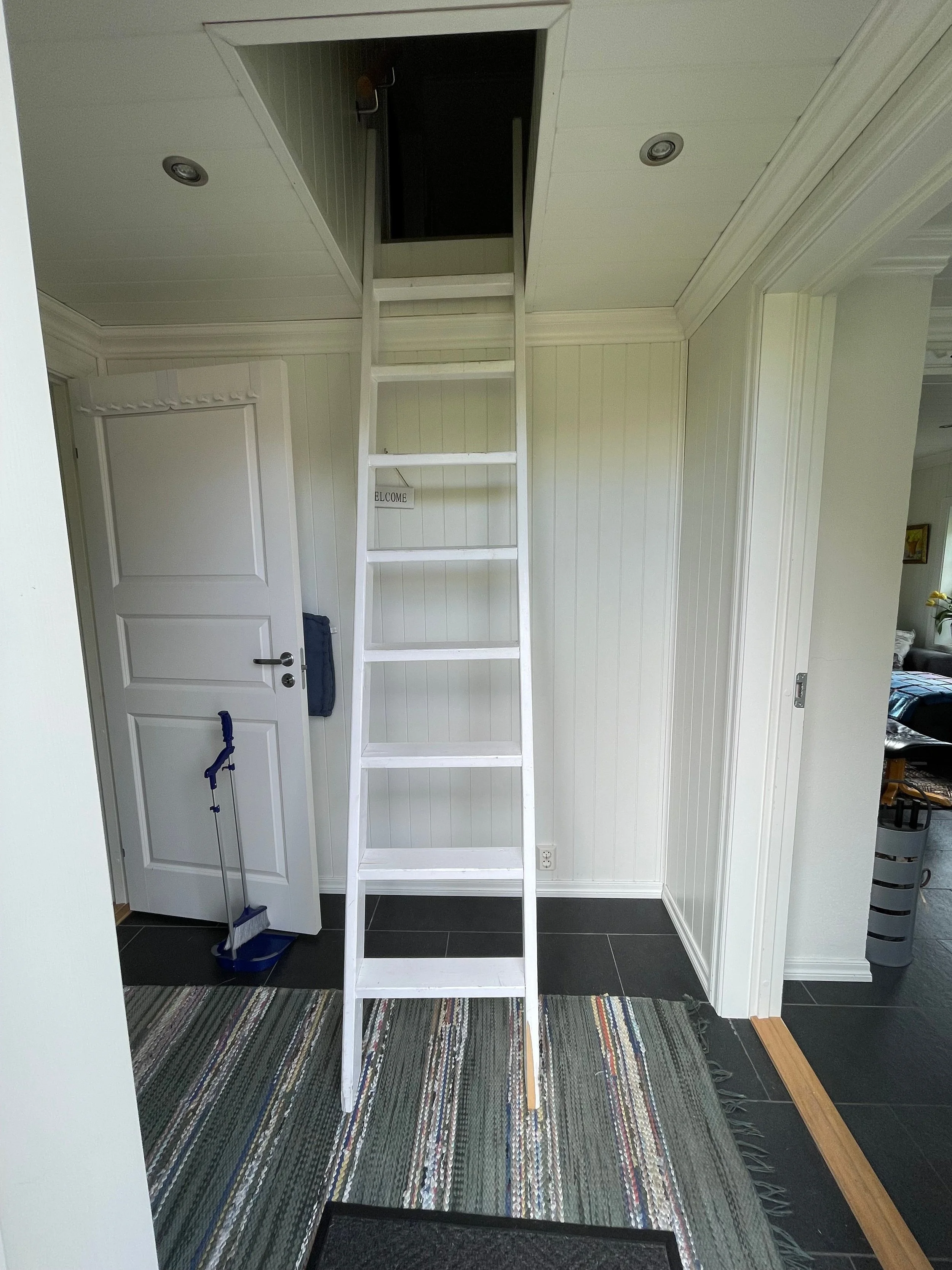 Ein weißer Holzleiter führt in einen dunklen Estrich. Die Raumdecke ist weiß und hat eingebaute Deckenleuchten. Ein weißer Türrahmen mit einem geschlossenen Türschild ist auf der linken Seite zu sehen. Es liegt ein Staubsauger und ein Besen in der Nä