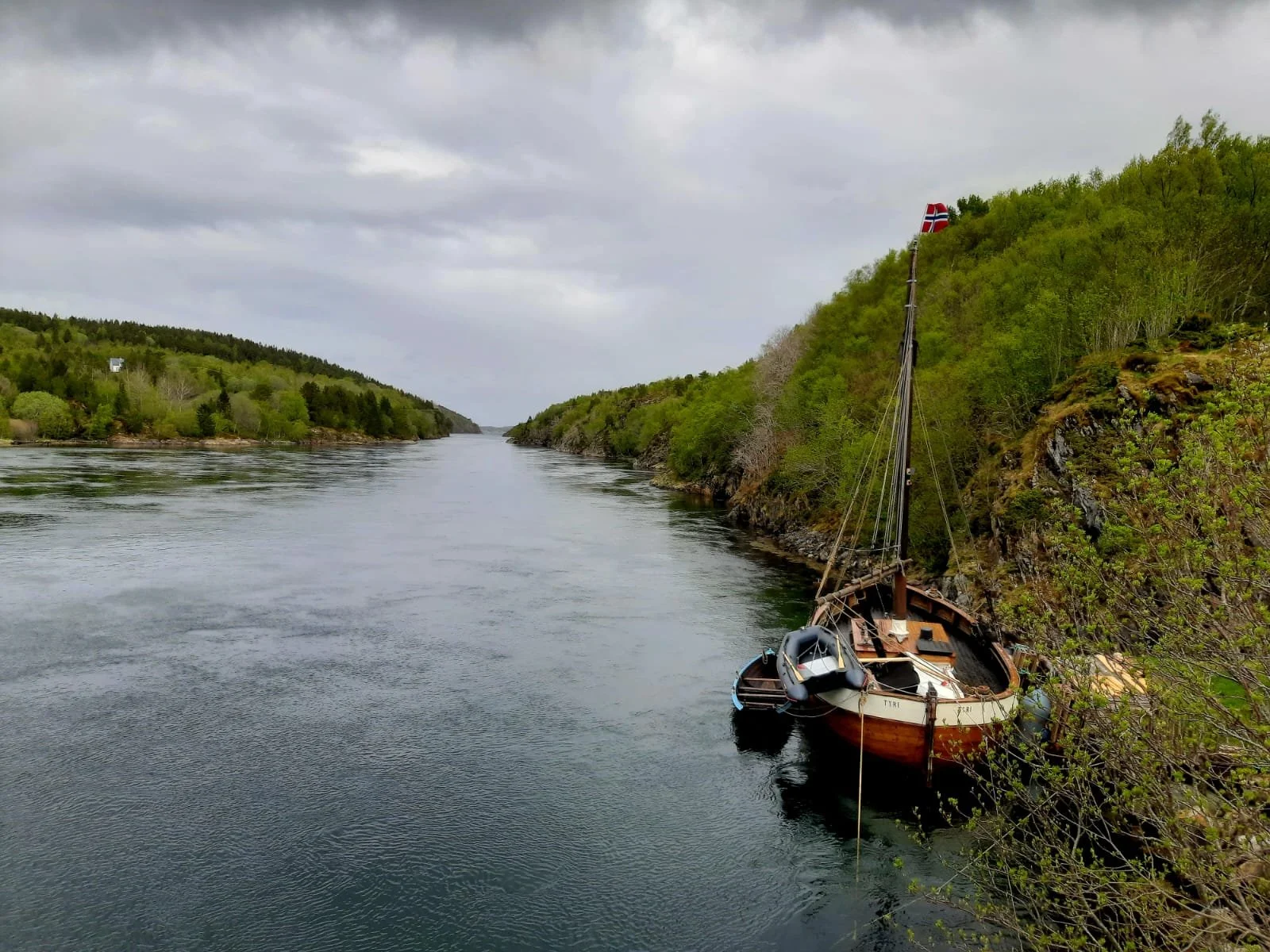 Ein Segelboot liegt an einem Flussufer, umgeben von grünen Bäumen, bei bewölktem Himmel.