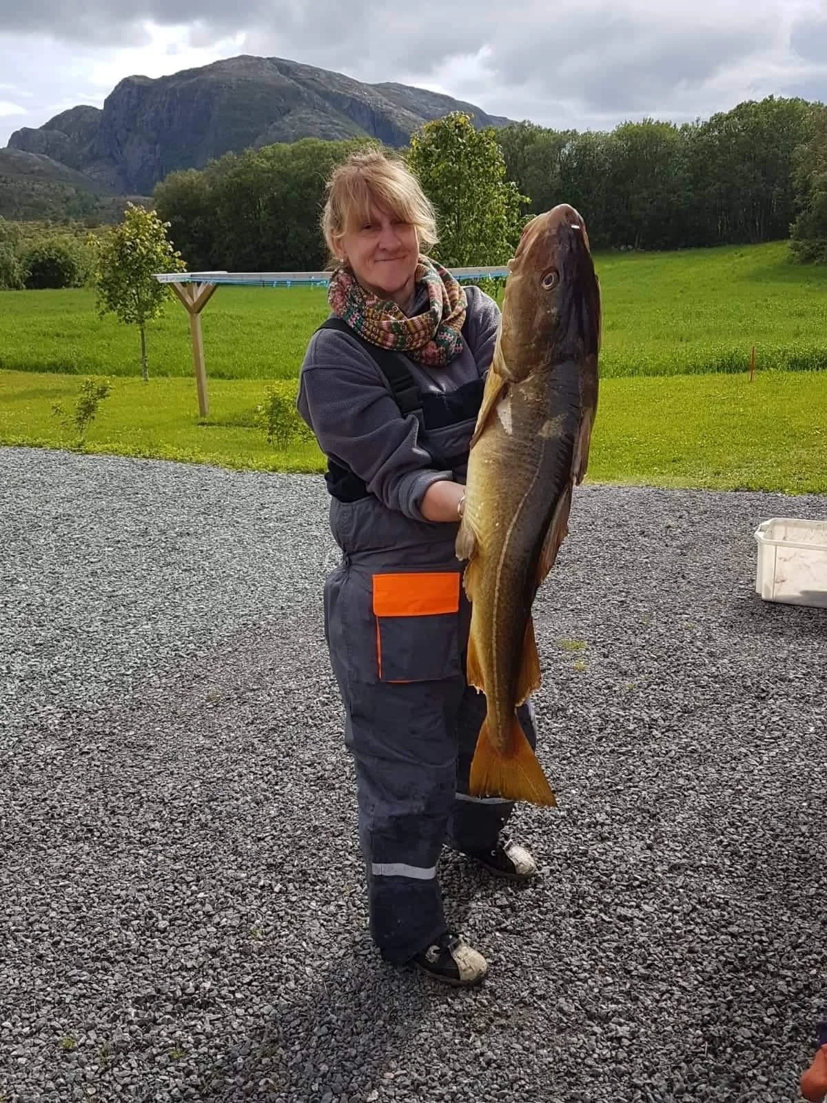 Frau hält große Fisch vor grünem Hintergrund mit Bäumen, Bergen und einer Wäscheleine.