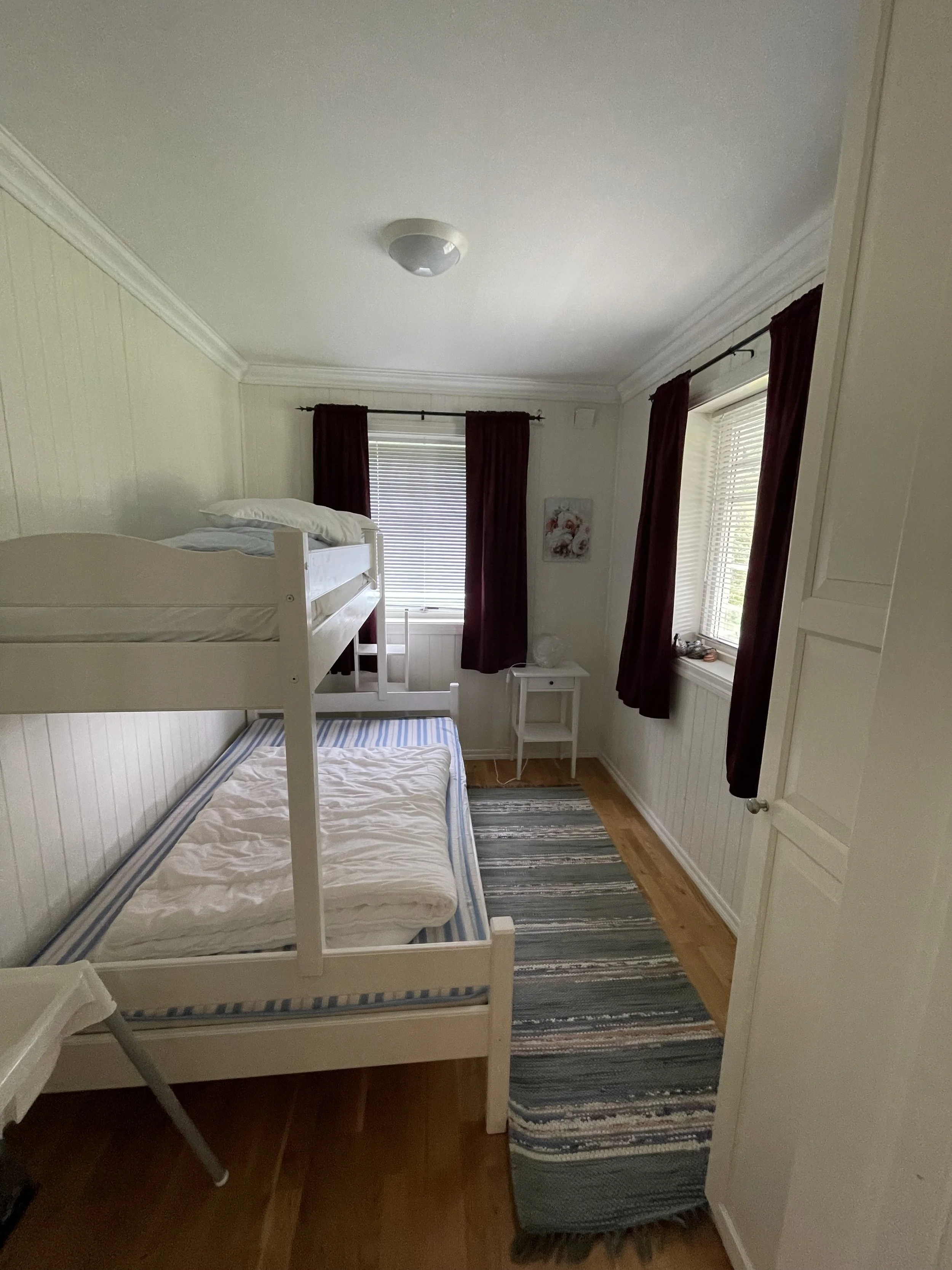 Ein Kinderzimmer mit einem weißen Hochbett, einem unteren Bett mit blauer Streifenmatratze und weißen Kissen, zwei Fenster mit roten Vorhängen, einem kleinen weißen Nachttisch, einer Wanddekoration und einem bunten langen Teppich auf Holzboden.