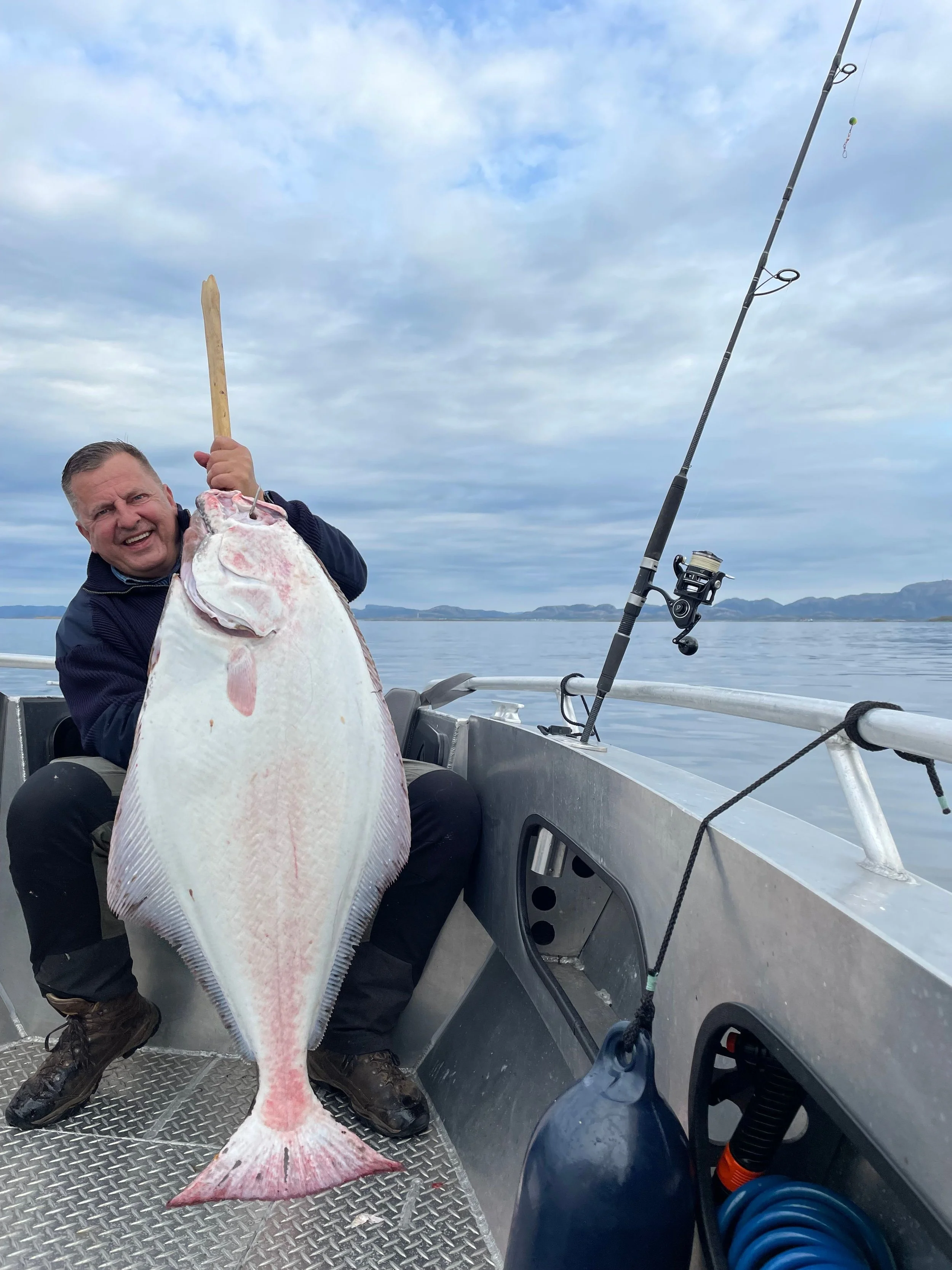 Ein lachender Mann hält auf einem Boot eine große, weiße Fisch mit einem Holzstich. Im Hintergrund ist das Wasser und ein bewölkter Himmel zu sehen.