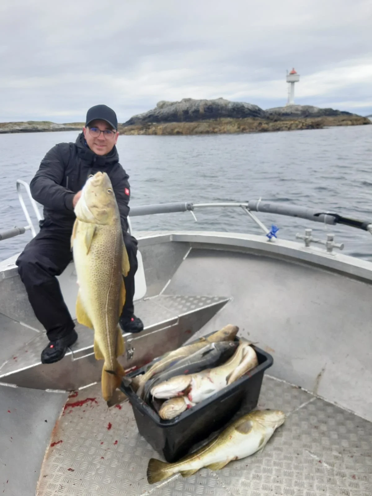 Ein Mann hält einen großen Fisch auf einem Boot. Es liegen weitere Fische in einem Behälter. Im Hintergrund ist eine Insel mit einem Leuchtturm zu sehen.